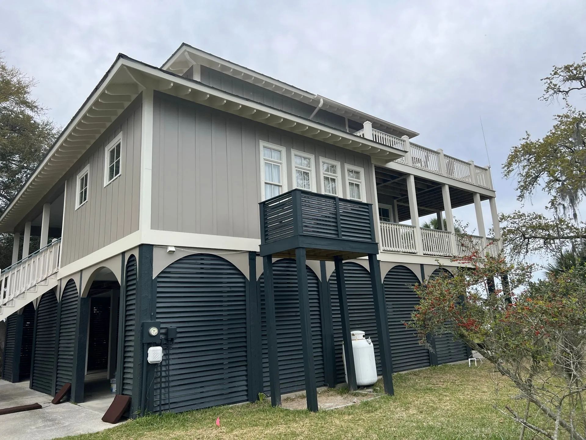 Raised coastal house with gray siding, dark shutters, and a wraparound porch on stilts