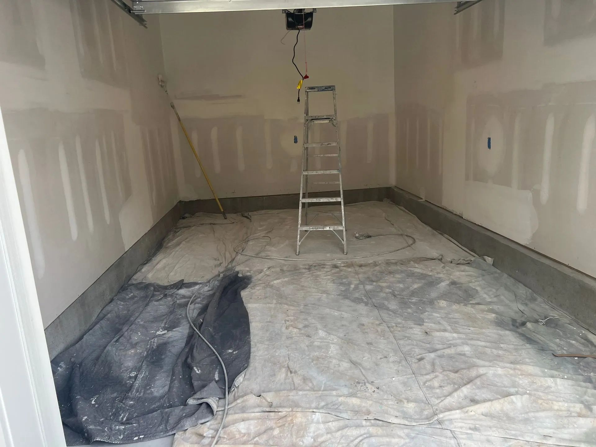 Partially renovated garage with drywall, tarp-covered floor, and a ladder under a ceiling light