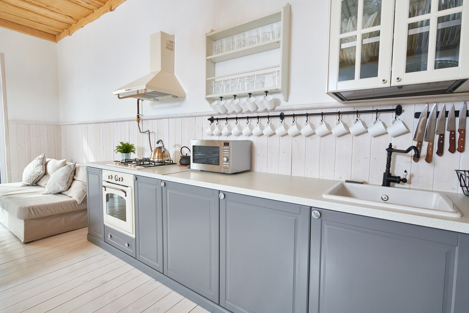 Gray and white kitchen with stove, sink, and cabinets. A microwave sits on the countertop with mugs hanging.