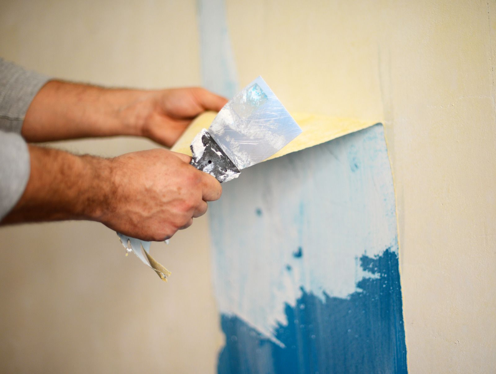 Person using a putty knife to scrape blue paint from a wall.
