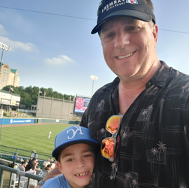 Smiling man and child in baseball caps at a ballpark, taking a selfie
