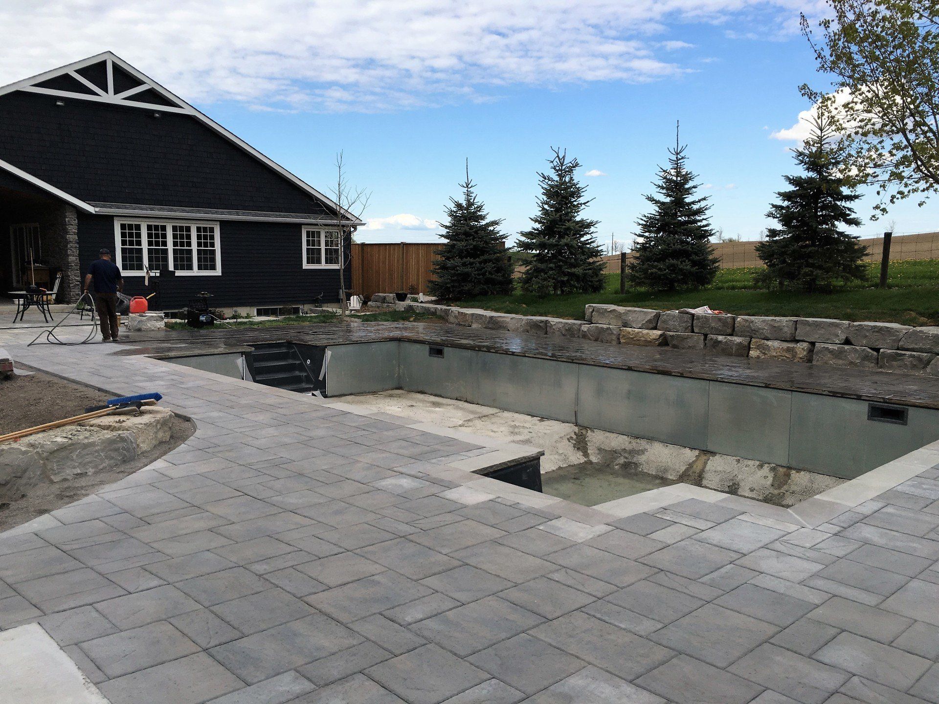 A concrete pool under construction with a paved patio, near a dark-colored house and evergreen trees.