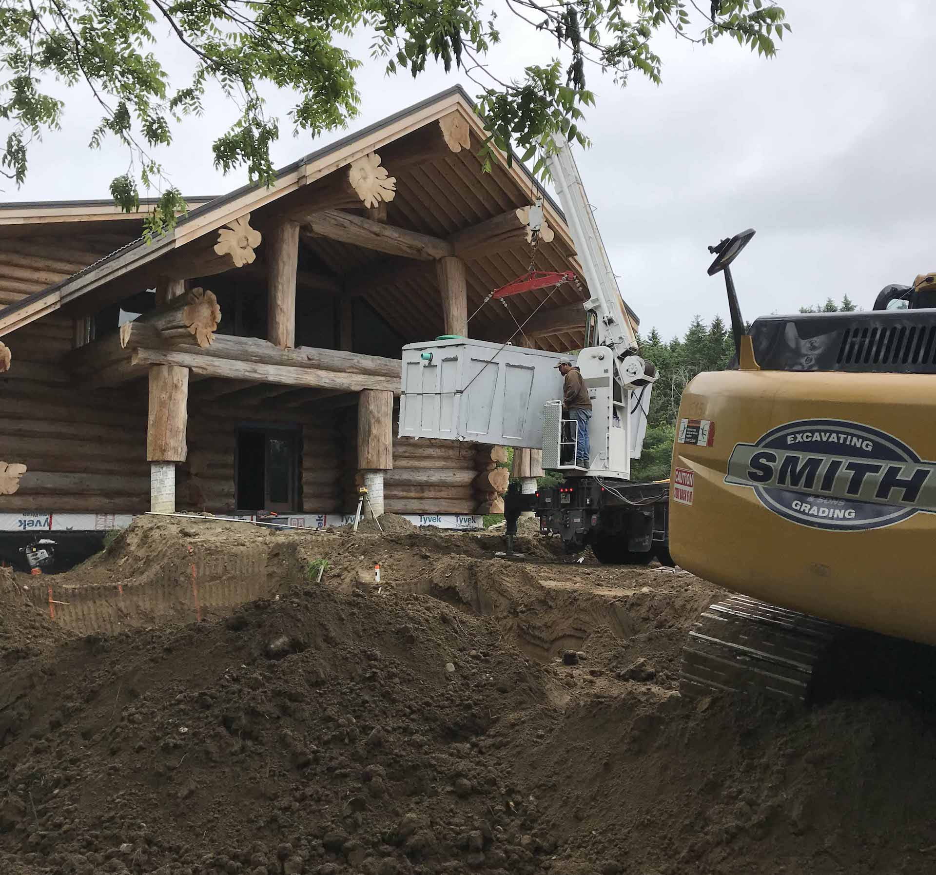 Construction site: excavator next to log cabin with a lift. Earth is piled up in front of the structure.