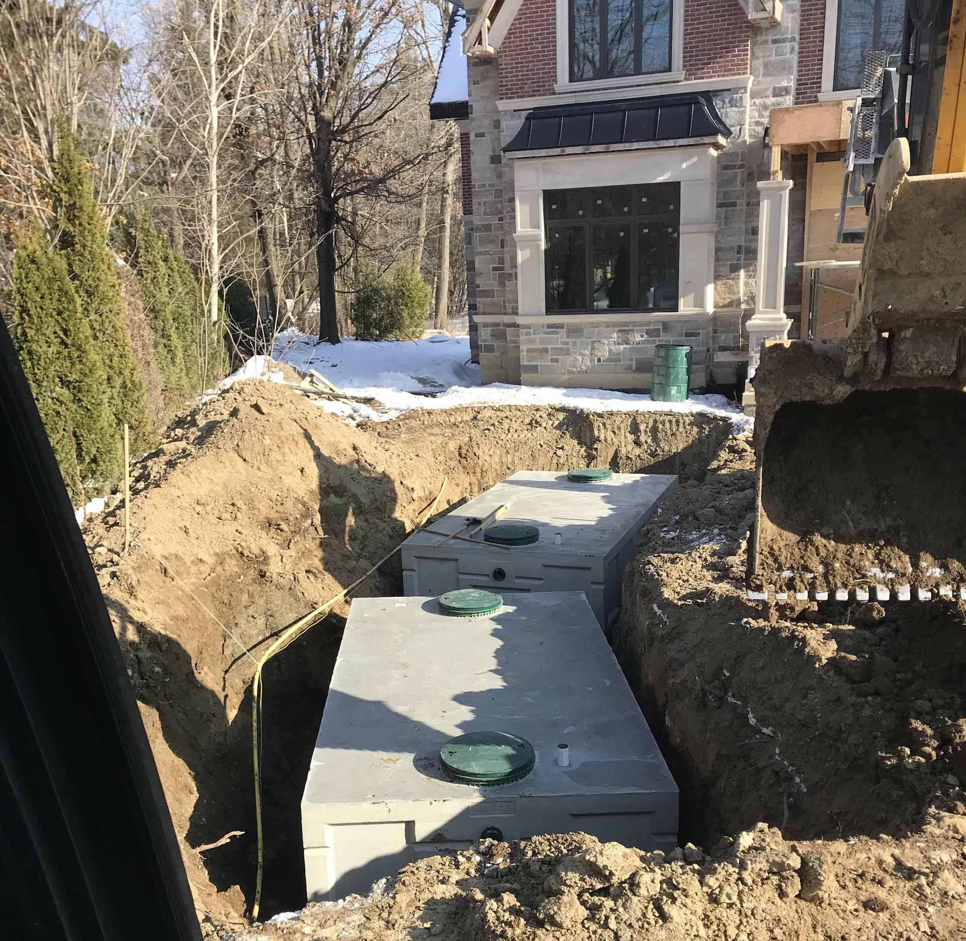 Excavated trench with two rectangular concrete septic tanks installed, next to a house under construction.