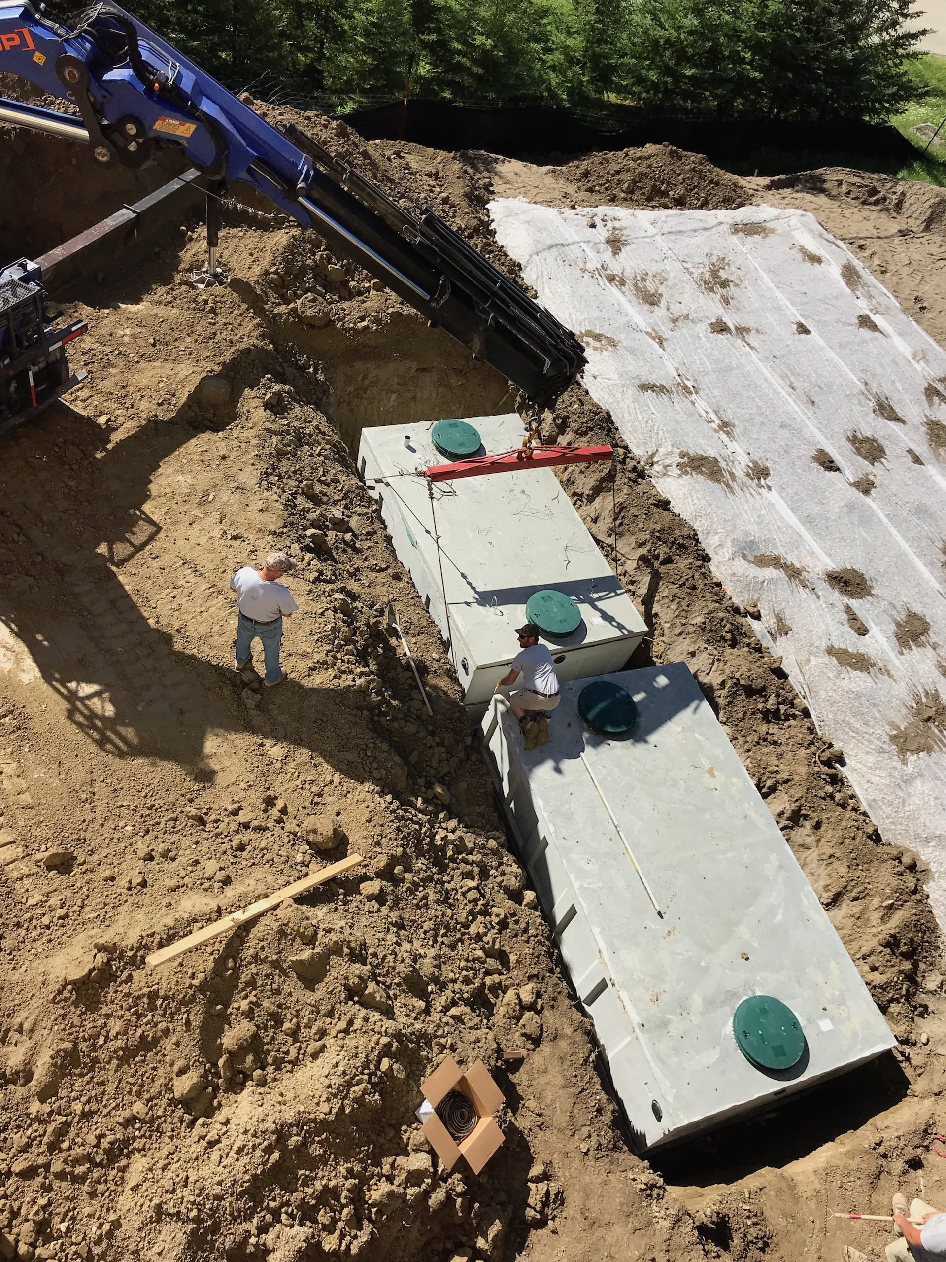 Excavator placing concrete septic tanks into a trench; workers nearby. Outdoors, sunny.