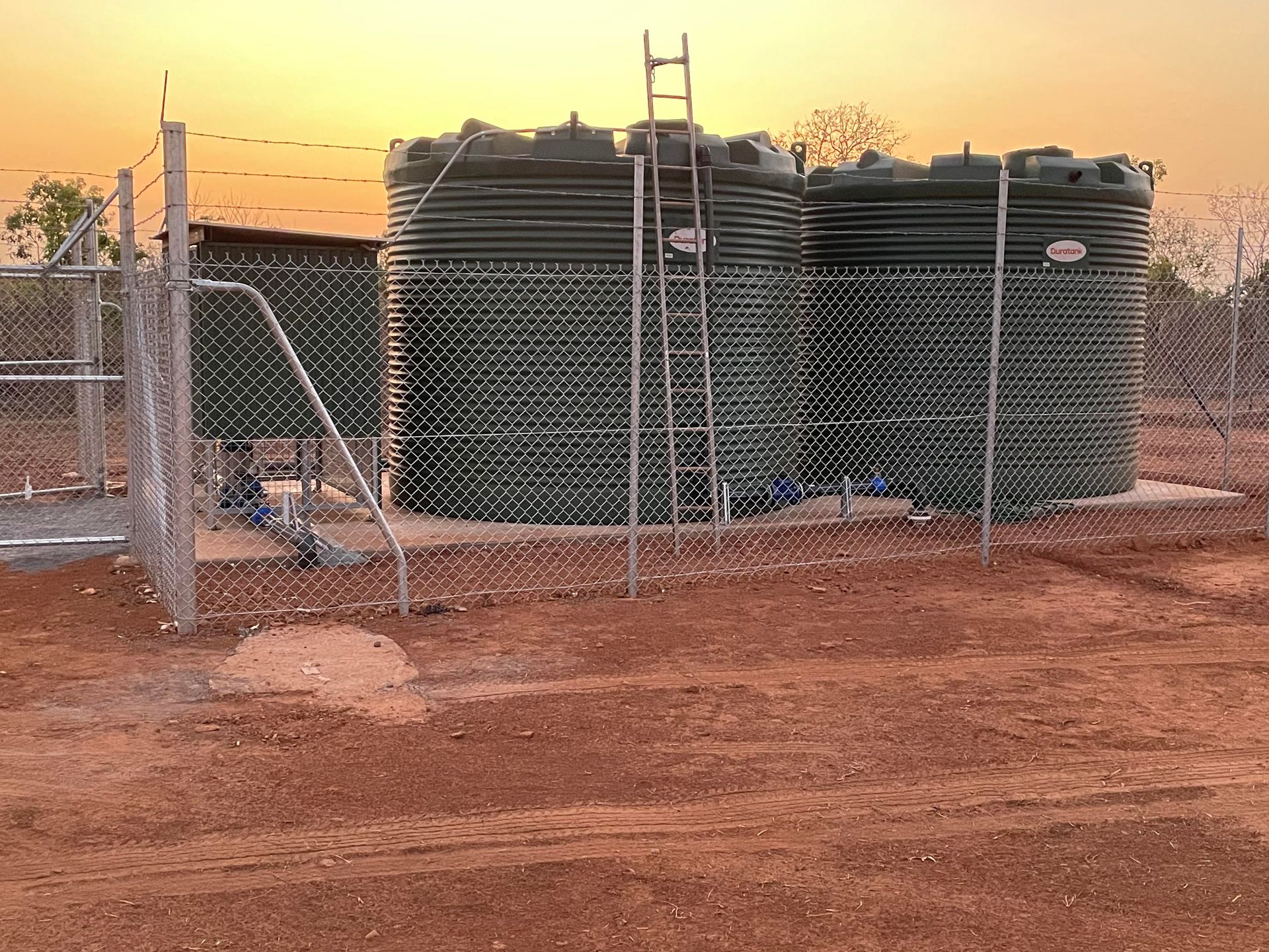 A fence surrounds a row of water tanks in a dirt field