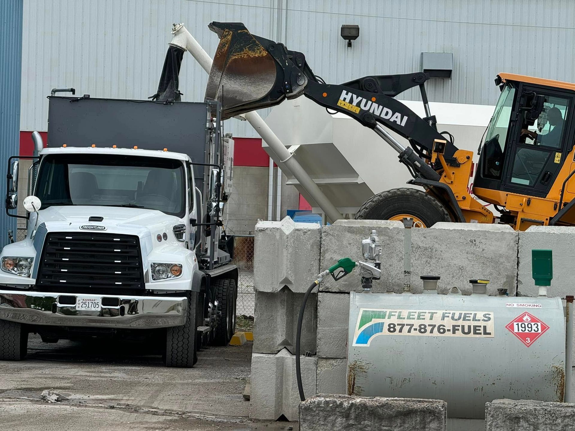 Excavator loading debris into a white dump truck at a construction site near concrete barriers.