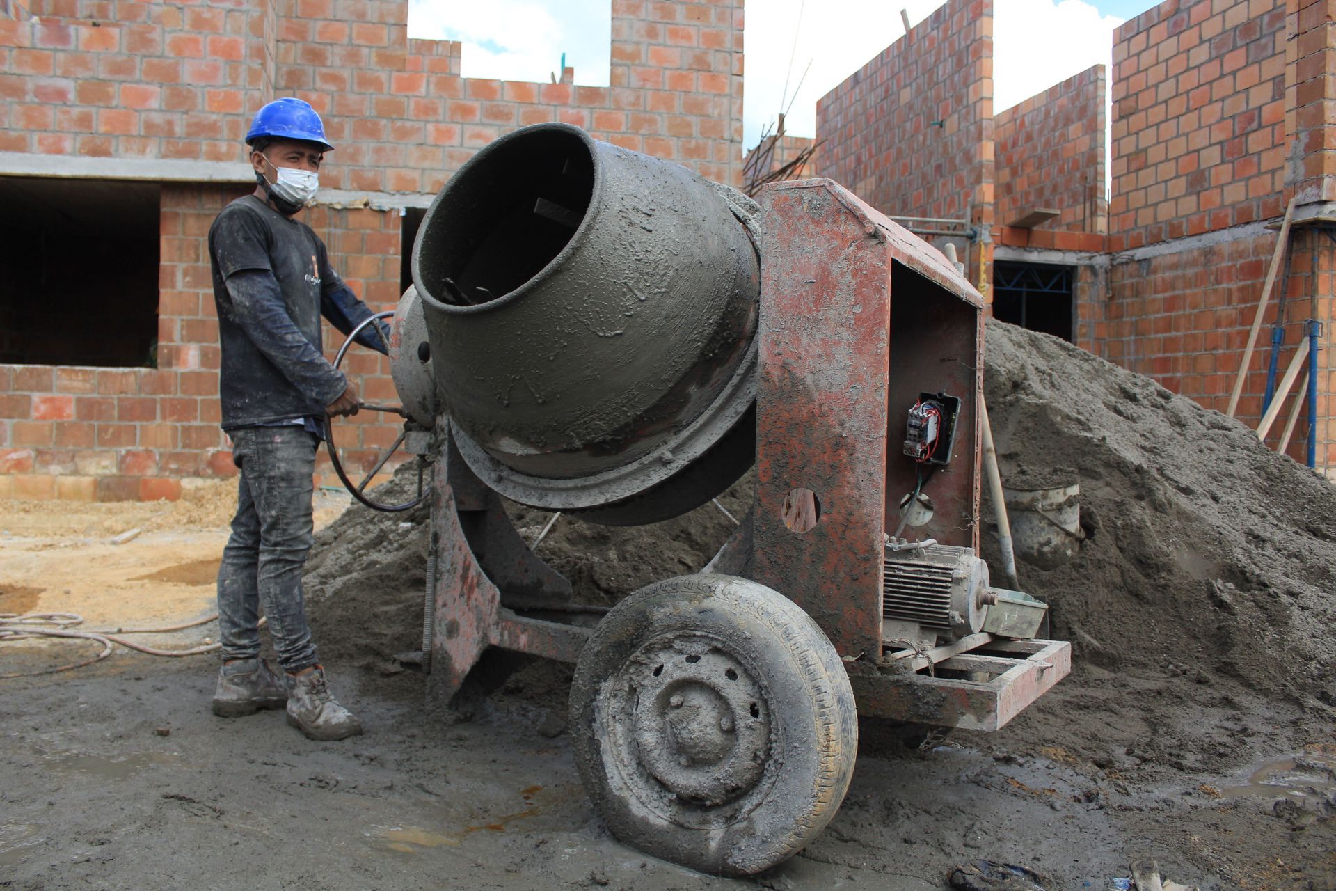 Construction worker beside a cement mixer at a brick building site, with piles of sand nearby