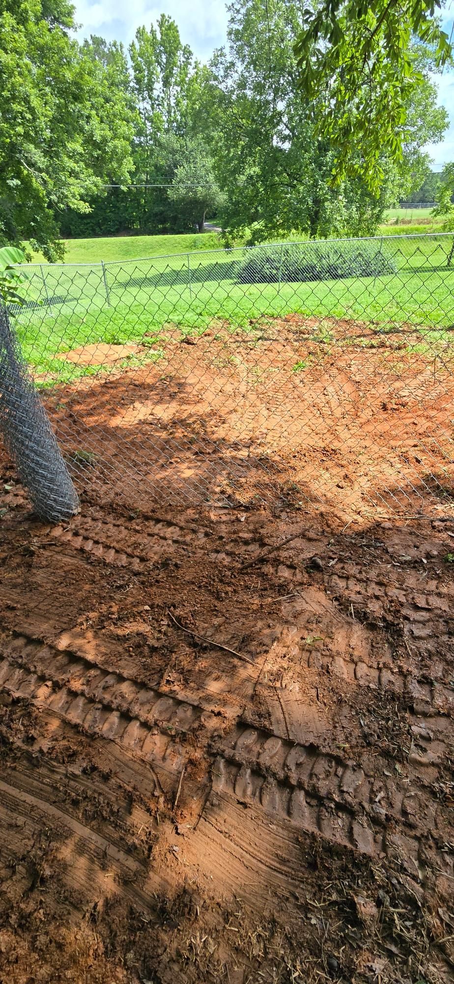 Muddy ground with tire tracks, a chain link fence, and a green, grassy field with trees in the background.