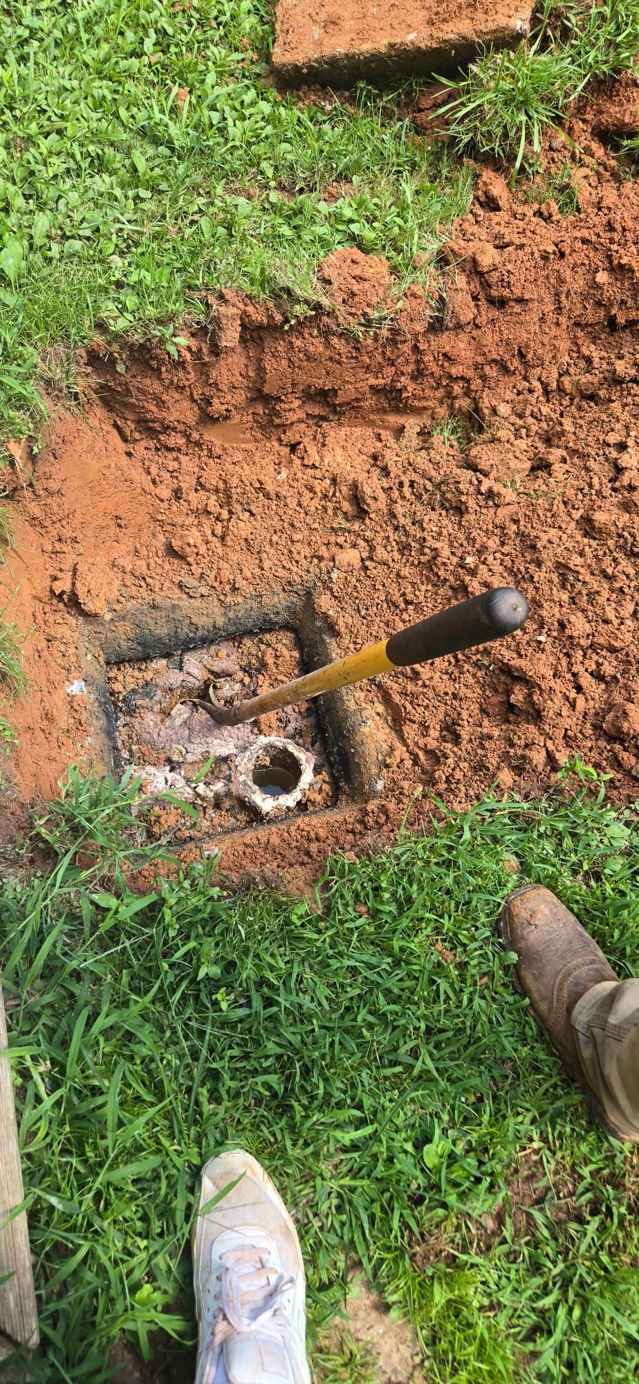 An open pit dug in the grass with a pipe exposed. A shovel and legs of a person are visible.