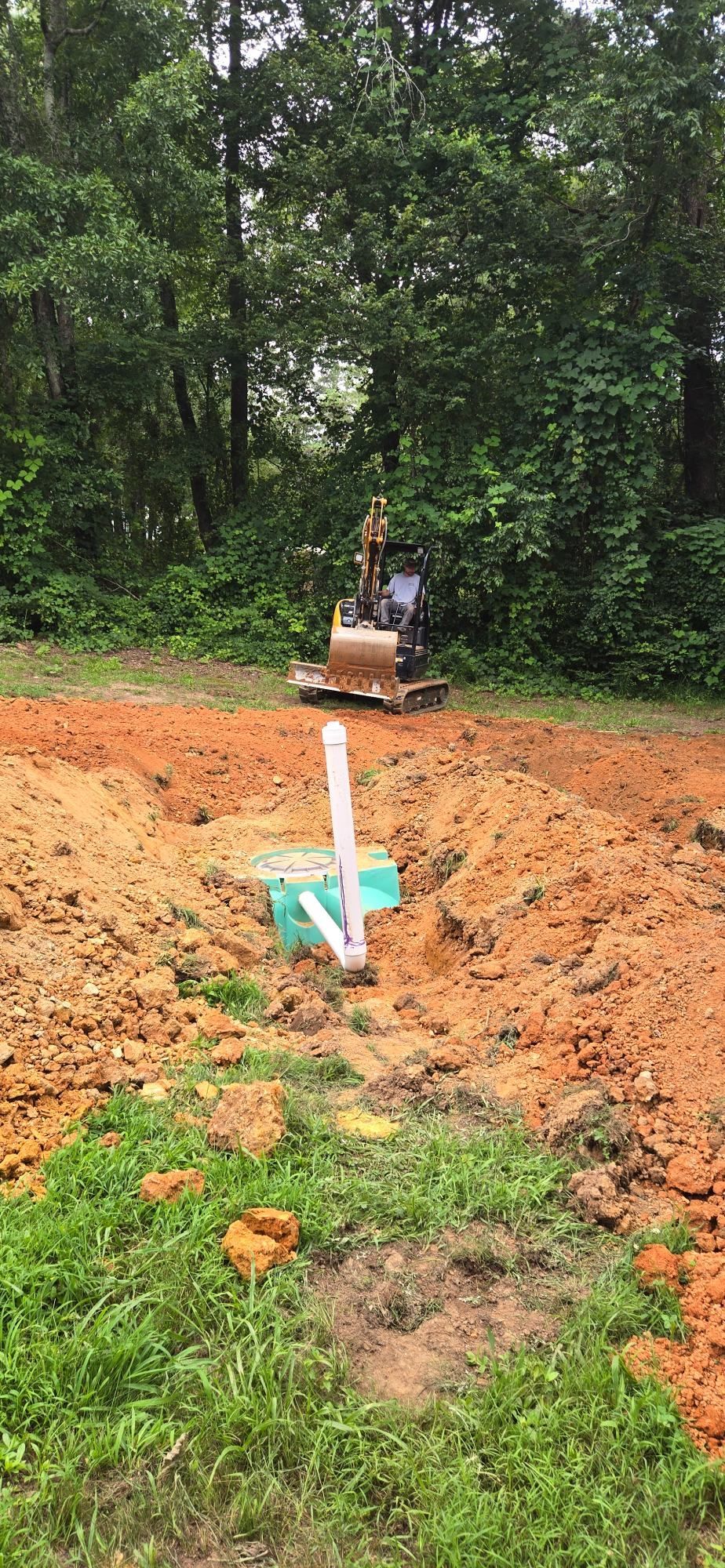Excavator digging a trench in a muddy backyard with a white pipe sticking out of the ground