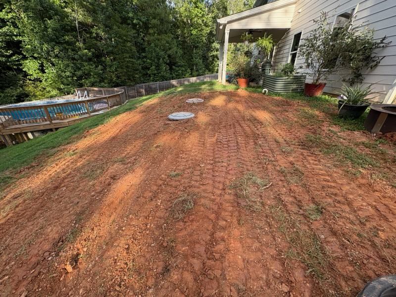 Freshly graded red dirt backyard beside a house, with potted plants and a porch in the background