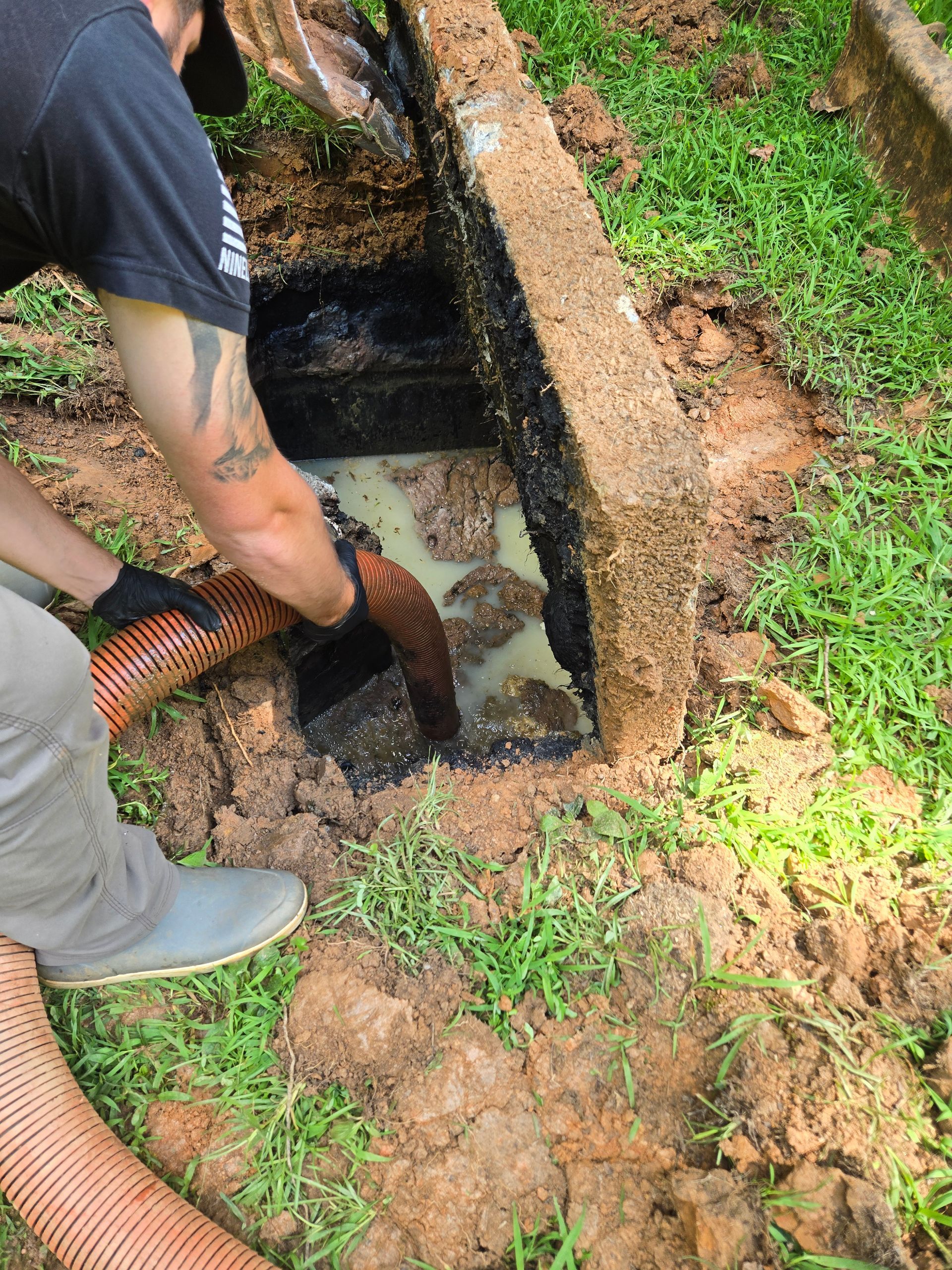 A person pumping sewage from an open septic tank, in a grassy yard. The sewage is a mix of light brown and dark sludge.