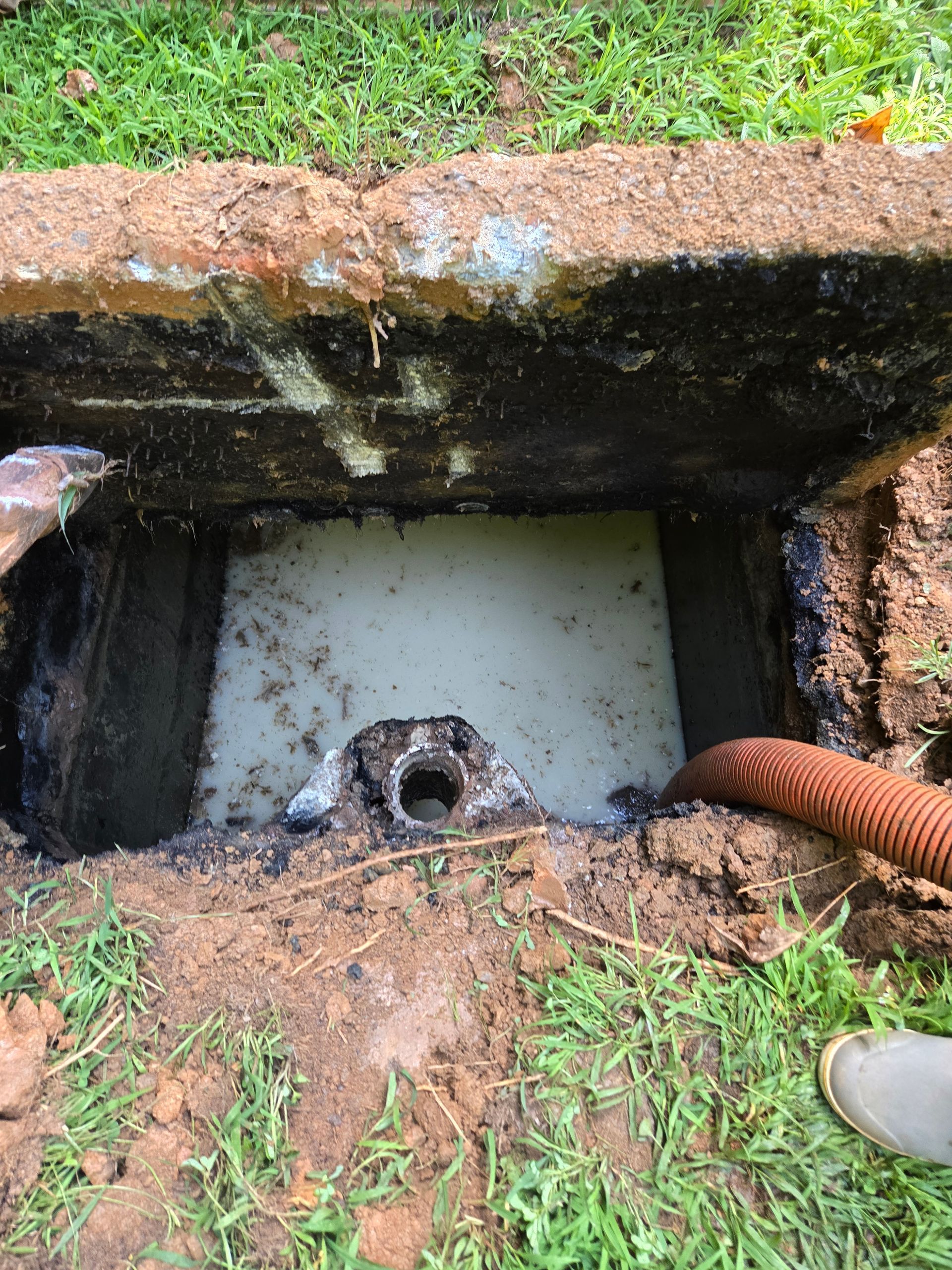 Open septic tank with visible inlet pipe, surrounded by dirt and a coiled orange hose. Concrete cover is above.