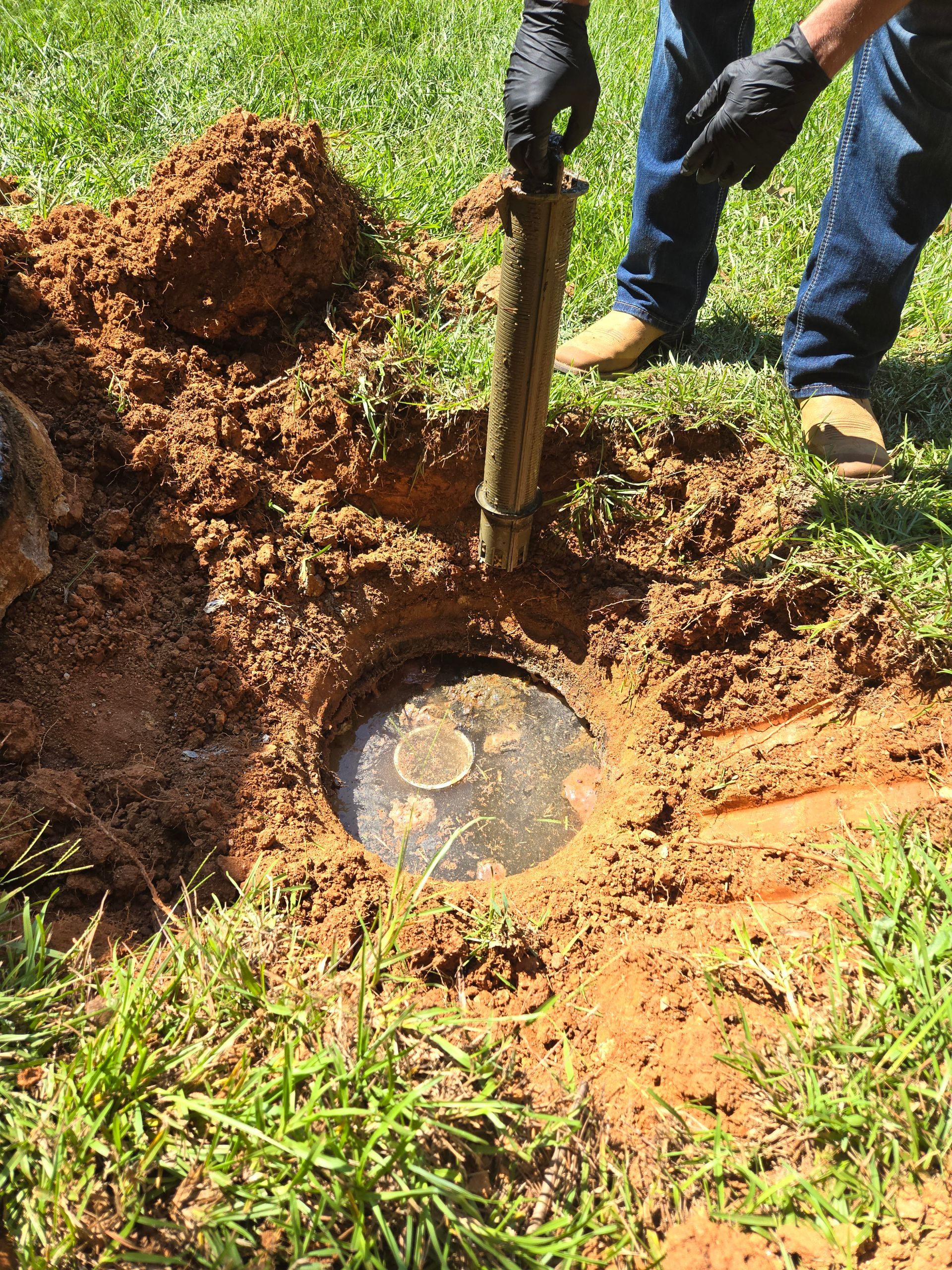 A person in blue jeans and gloves is inspecting a buried tank, possibly a septic system, in a lawn.