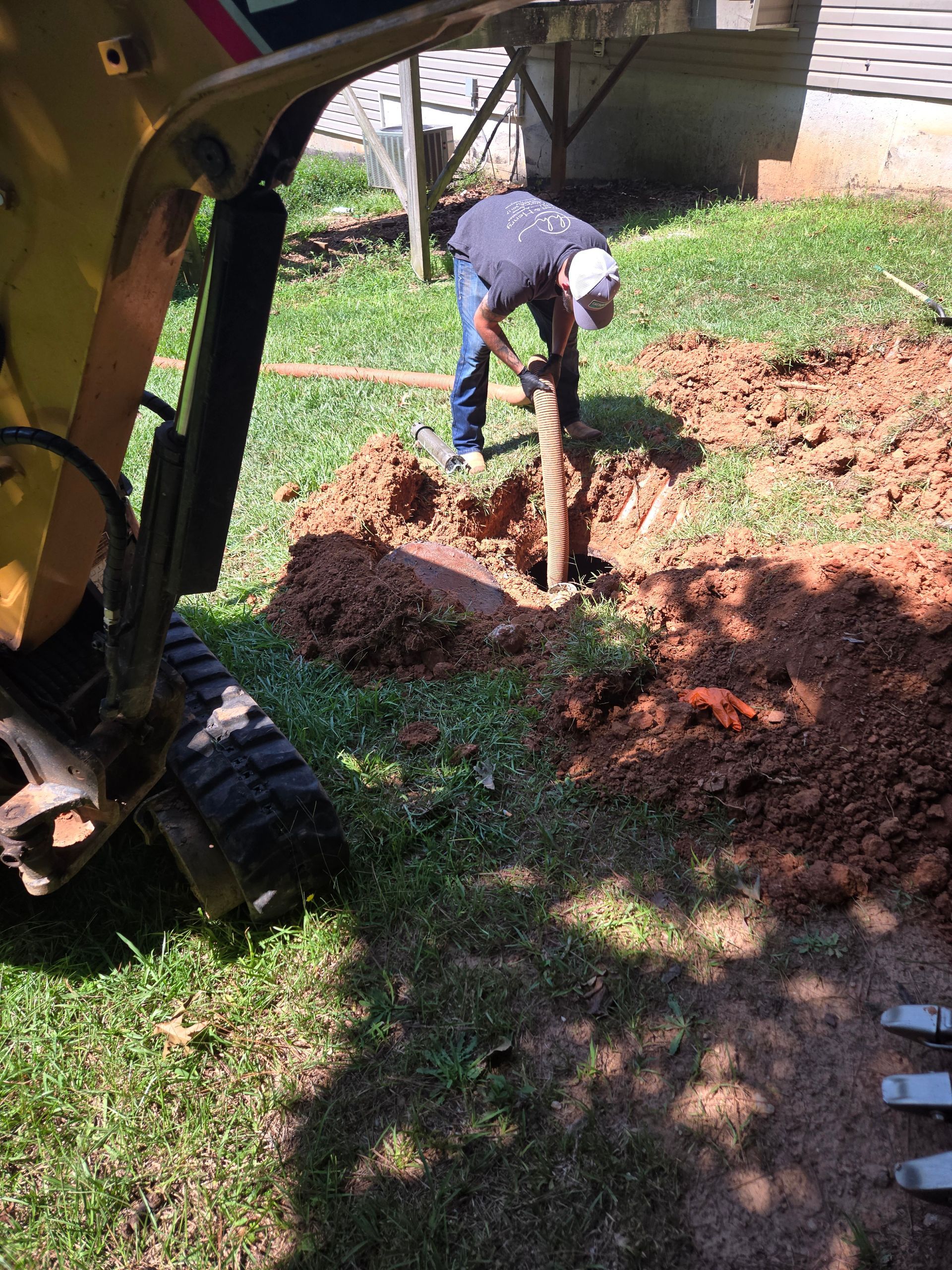 Man digging a hole in a grassy yard with a yellow construction vehicle nearby.