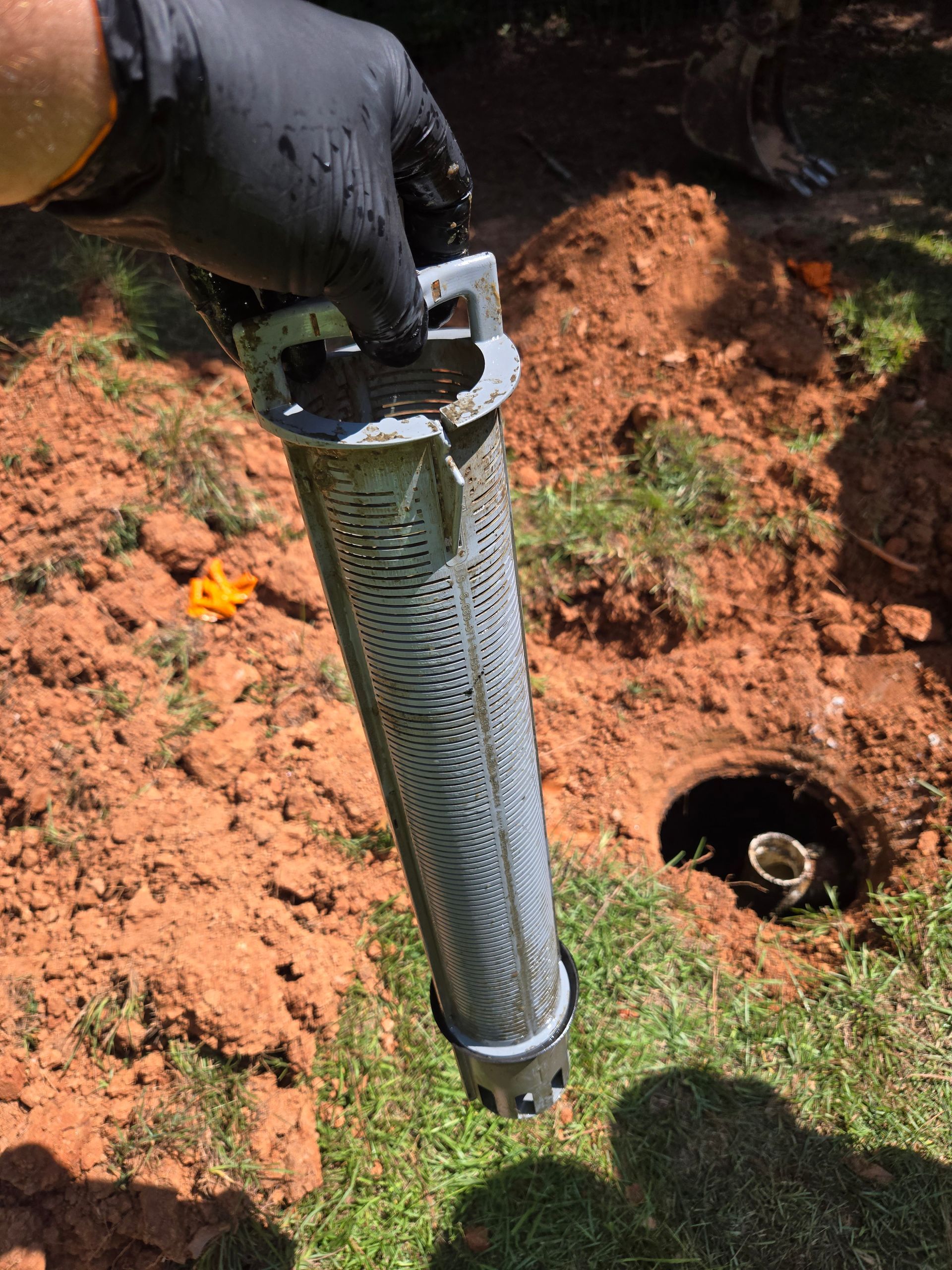 A person in a black glove holds a cylindrical well screen near an excavated hole in the ground.