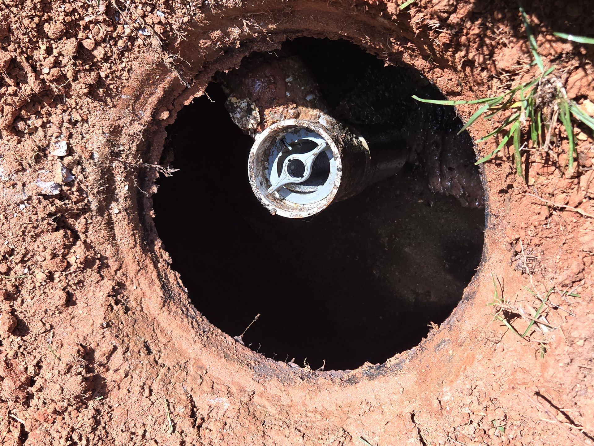 A deep hole in red dirt reveals a plumbing cleanout with a white plastic pipe.