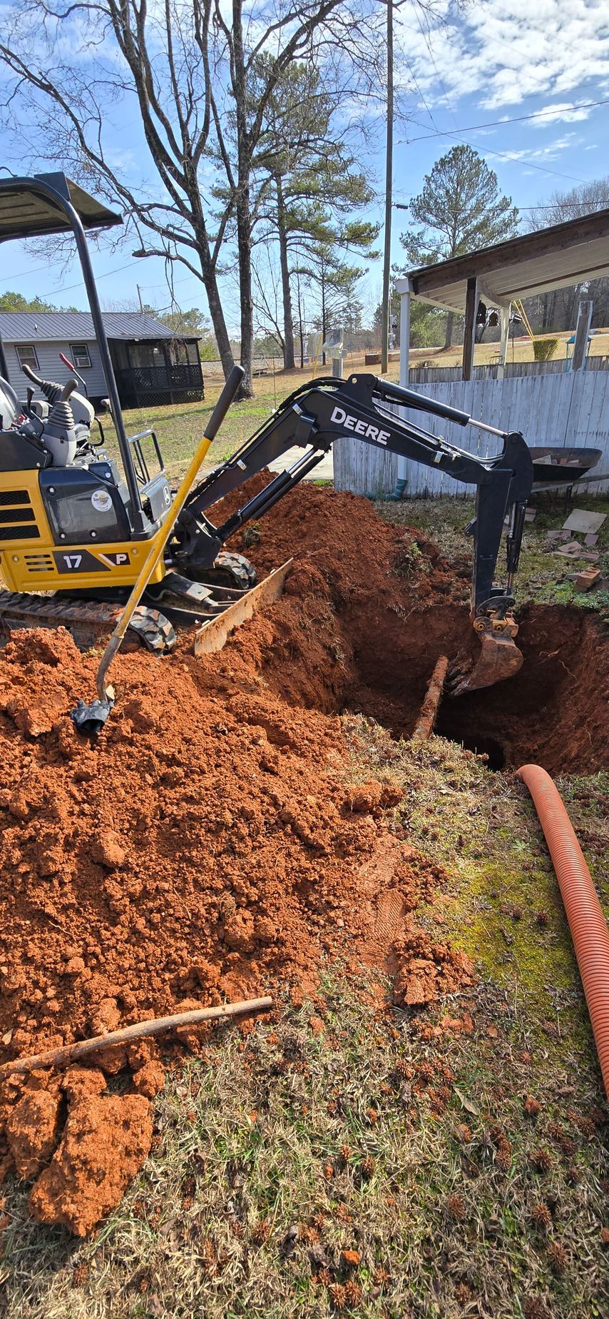 A small excavator digging in reddish-brown earth next to a patch of grass. A long, orange pipe lies in the grass.