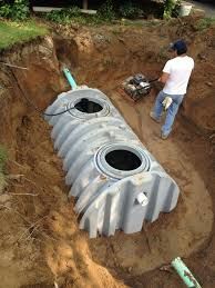 A person installs a septic tank in a freshly dug pit. The tank is gray, and the person is wearing a white shirt and jeans.