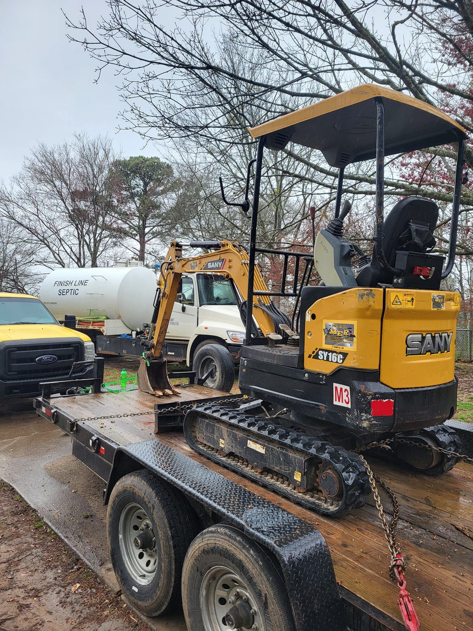 Yellow mini excavator on a trailer, with a white tank truck behind it. Cloudy day.