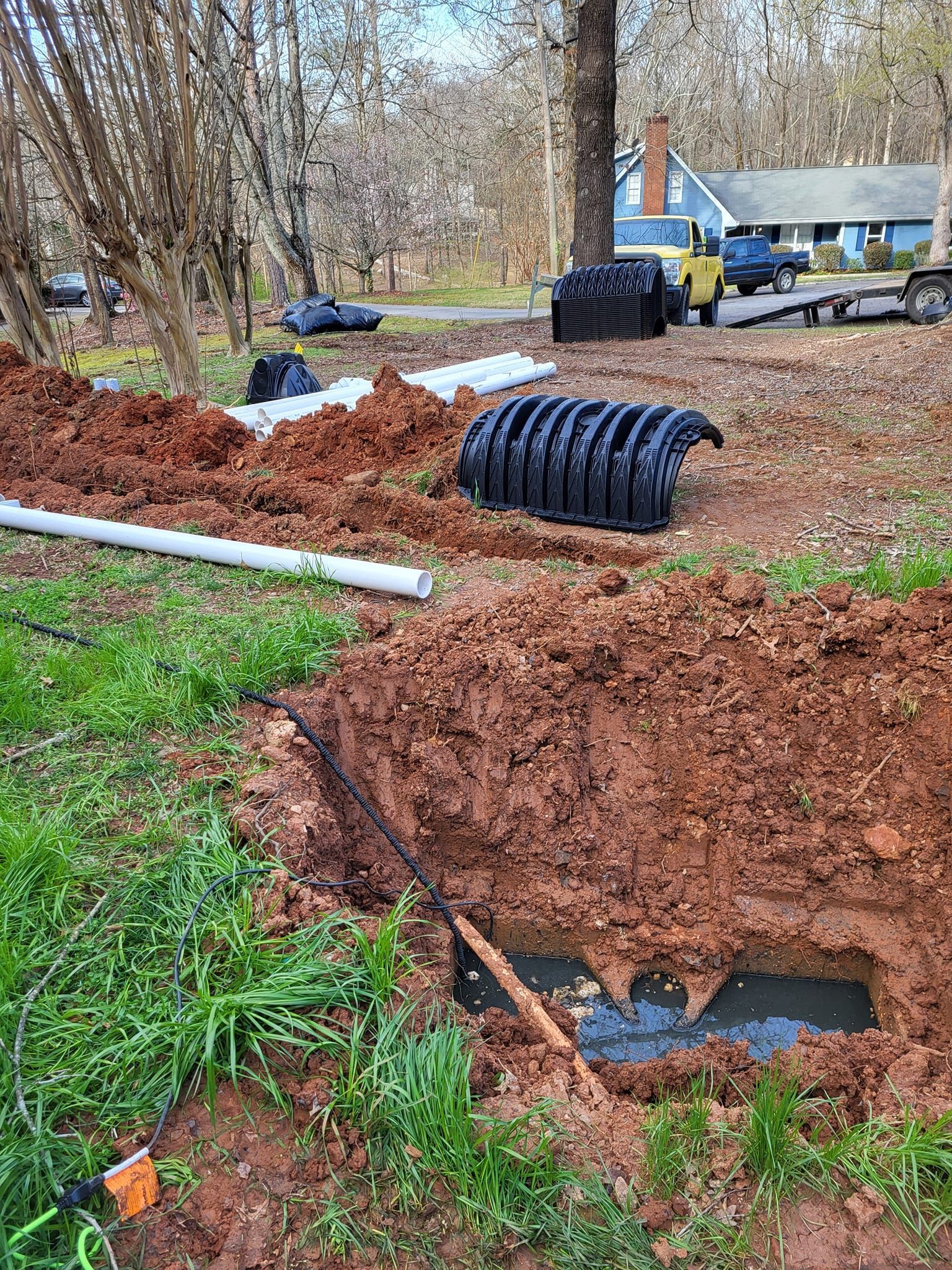 Excavation site in a yard with exposed pipes and a dark, corrugated structure.  Red-brown soil surrounds the hole.