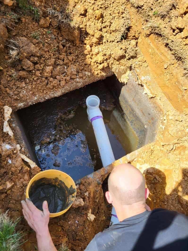 Man working on a buried pipe in a hole. A white PVC pipe with a purple band is submerged in water.