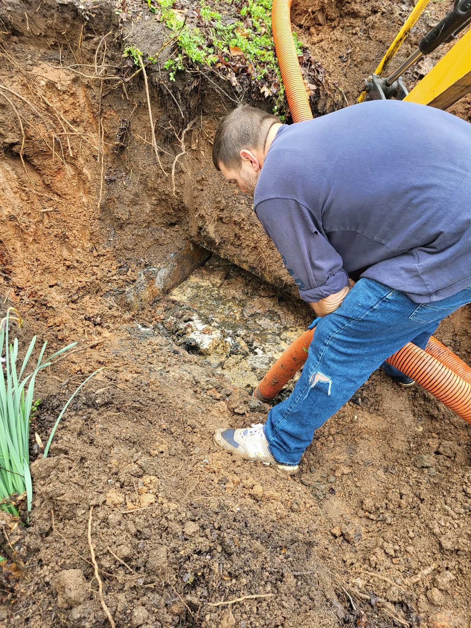 Man in blue shirt and jeans inspecting a hole in the ground near an orange hose; dirt surrounds him.
