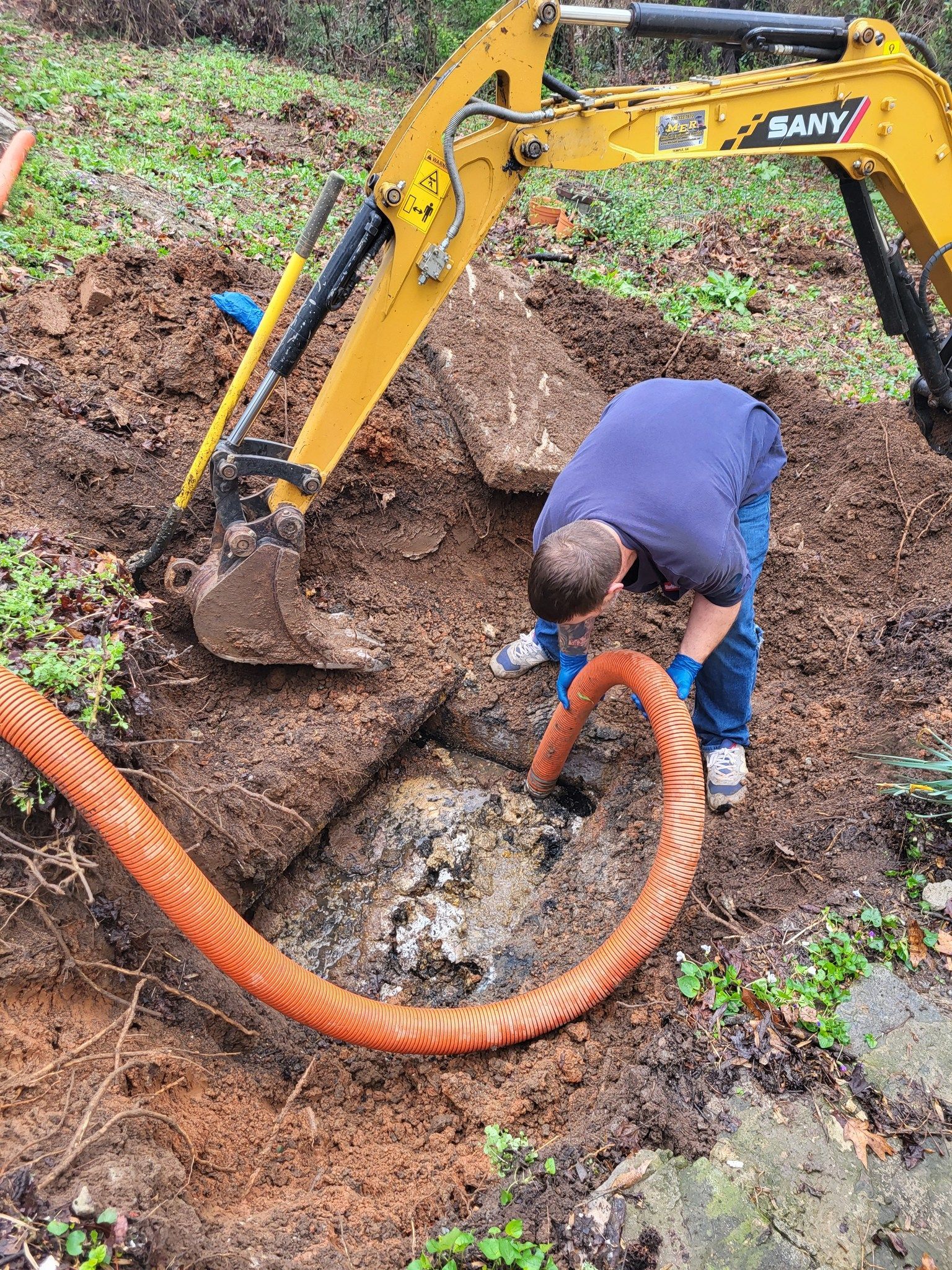 Man in blue shirt and jeans using a vacuum hose in a muddy excavation pit near a yellow excavator.