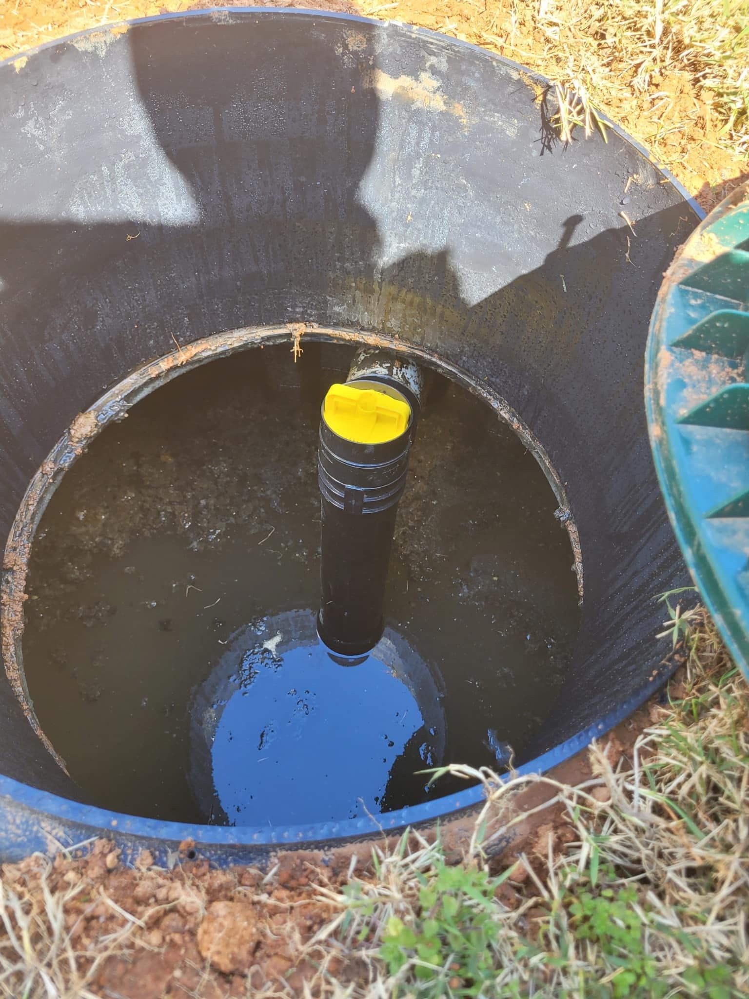 Open septic tank with standing water, a black pipe with a yellow cap, and surrounding dirt and grass.