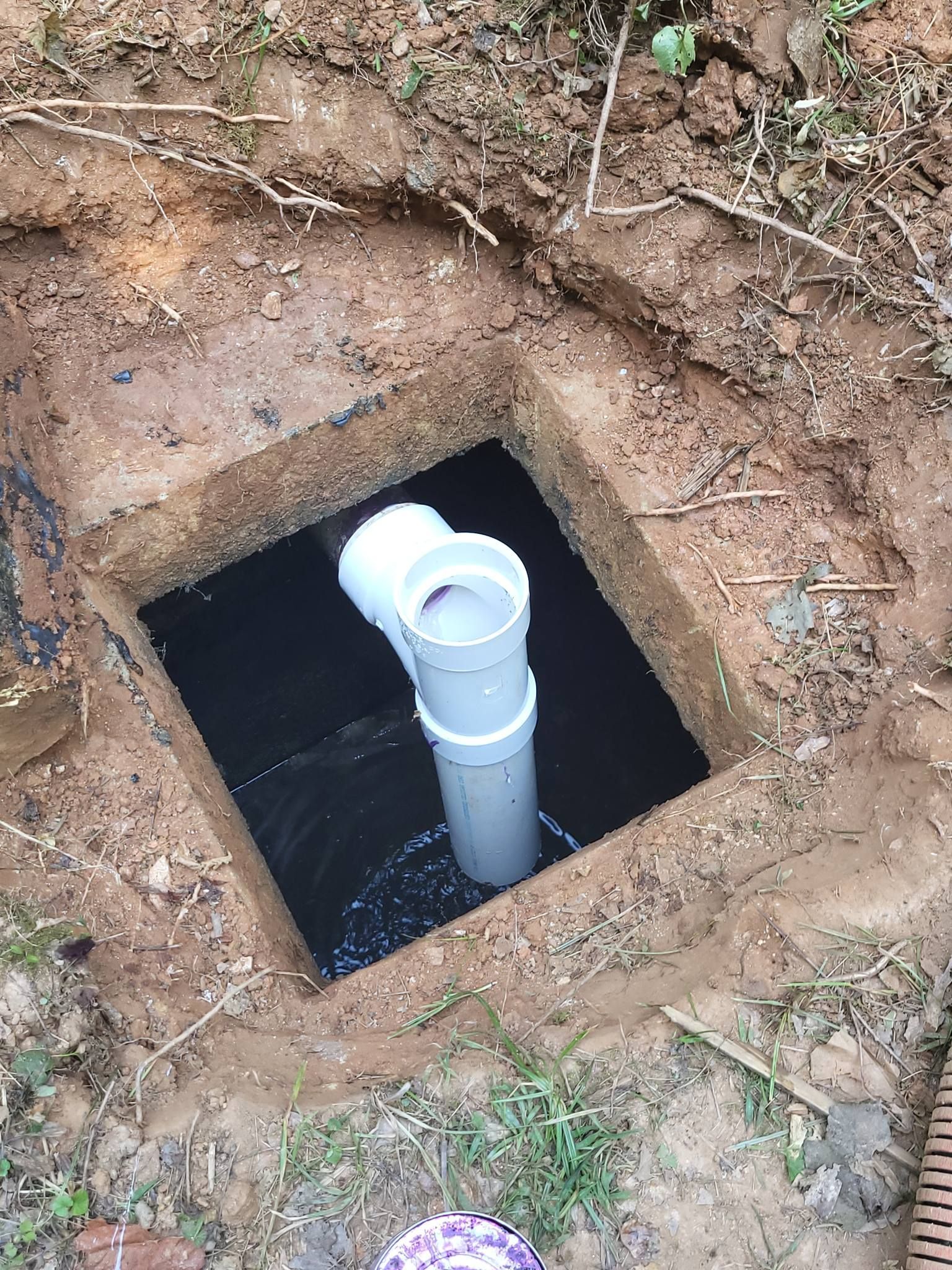 Open square pit in brown earth with a white PVC pipe and T-connector leading down into a dark interior.