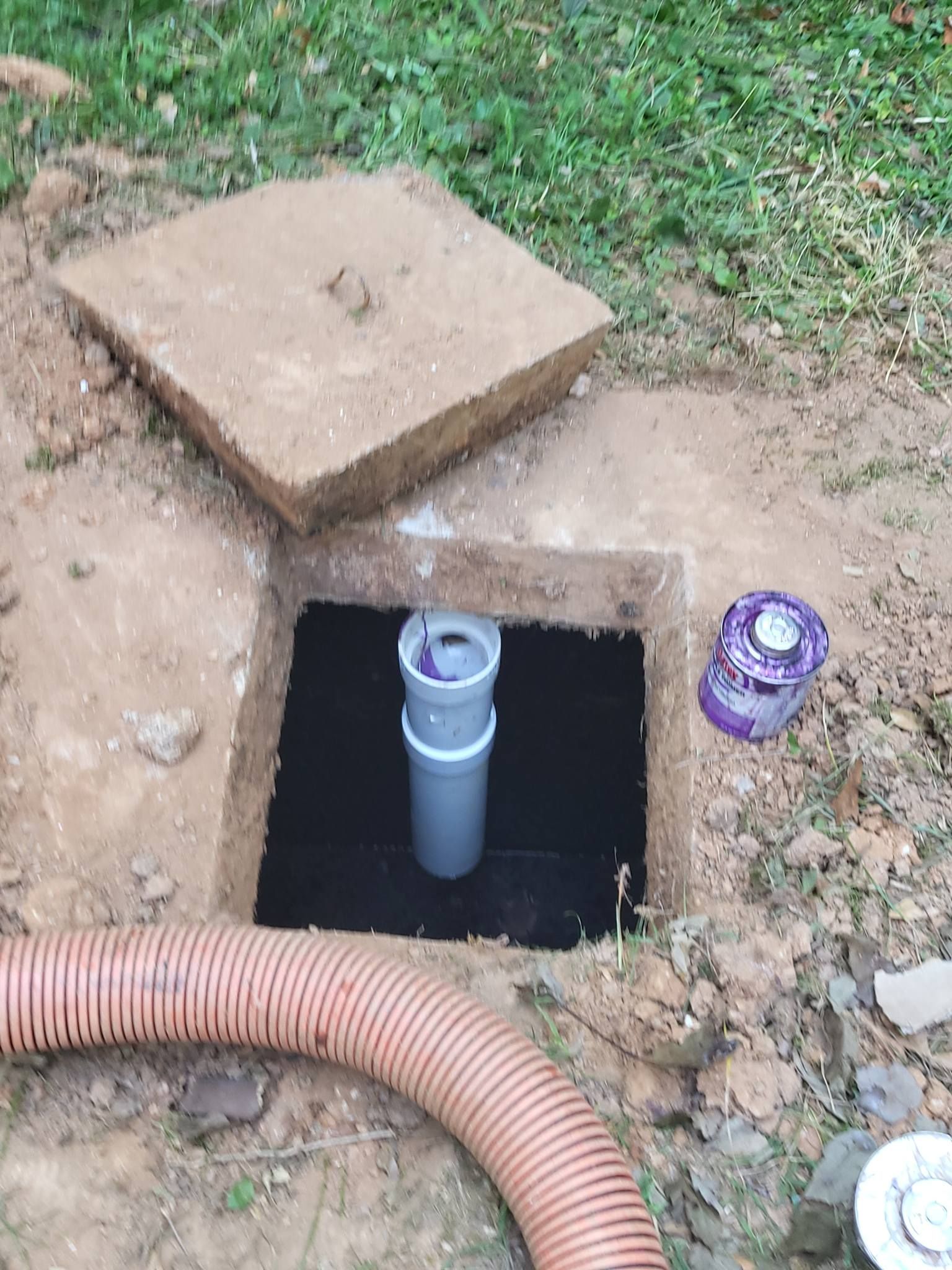 Open access point in a yard containing a pipe and corrugated hose. A concrete cover and small paint can are nearby.