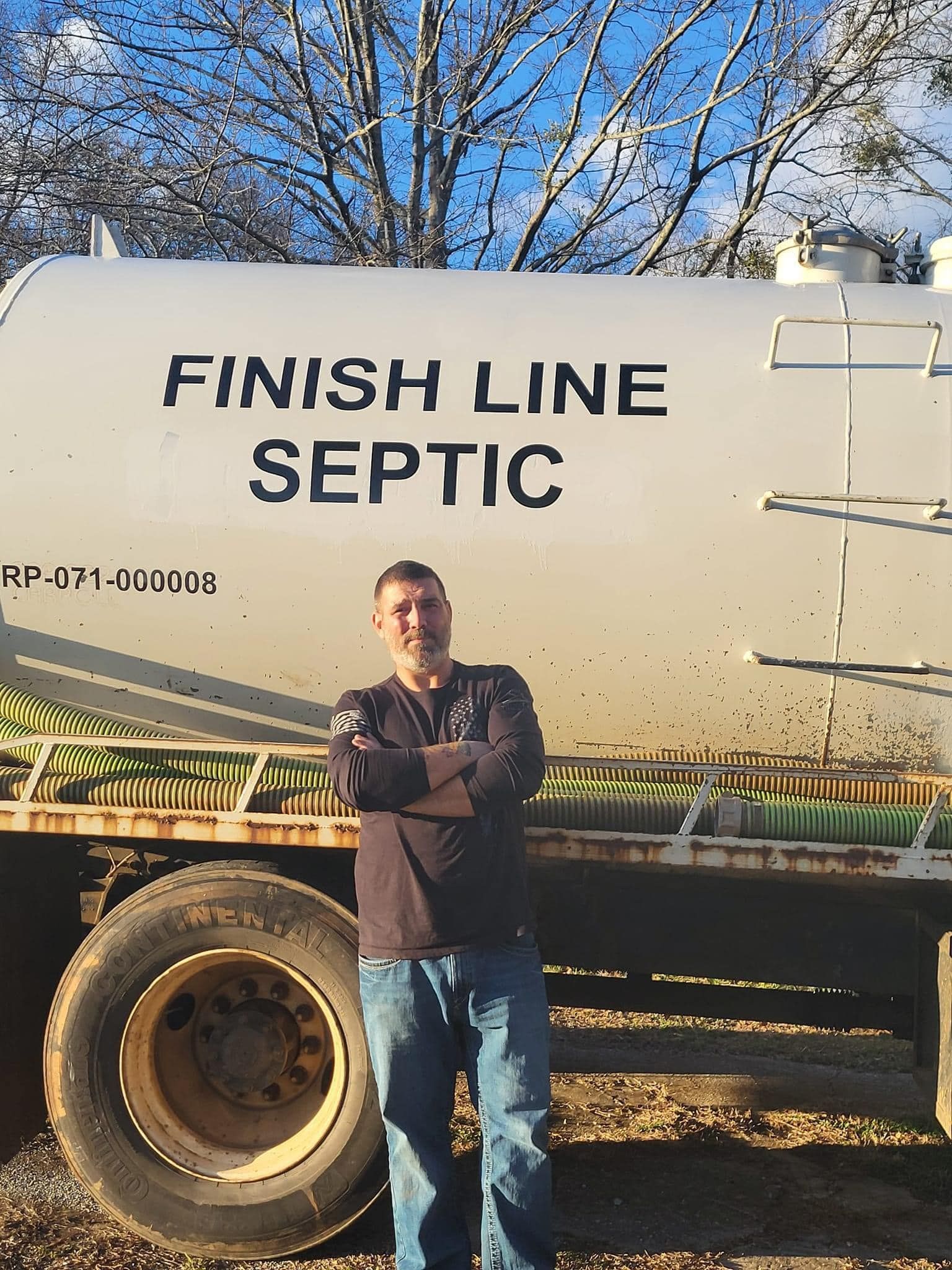 A man stands in front of a septic tank truck, his arms crossed. The truck is white with