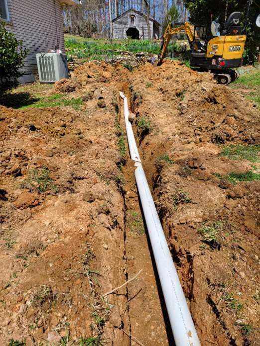 A trench dug in a yard with a white pipe running through it, an excavator in the background.