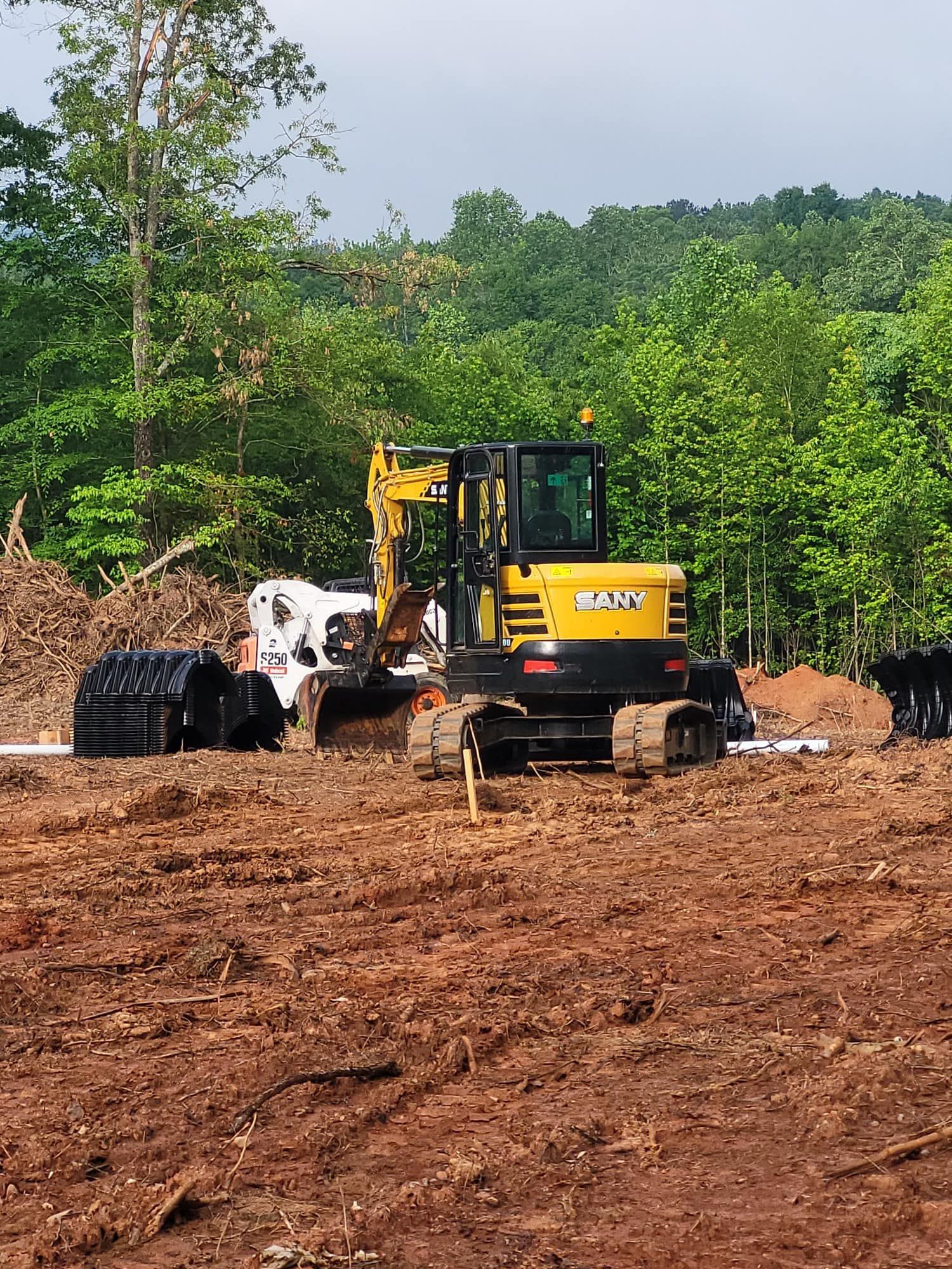 A yellow SANY excavator at a construction site, clearing land with a backdrop of trees.