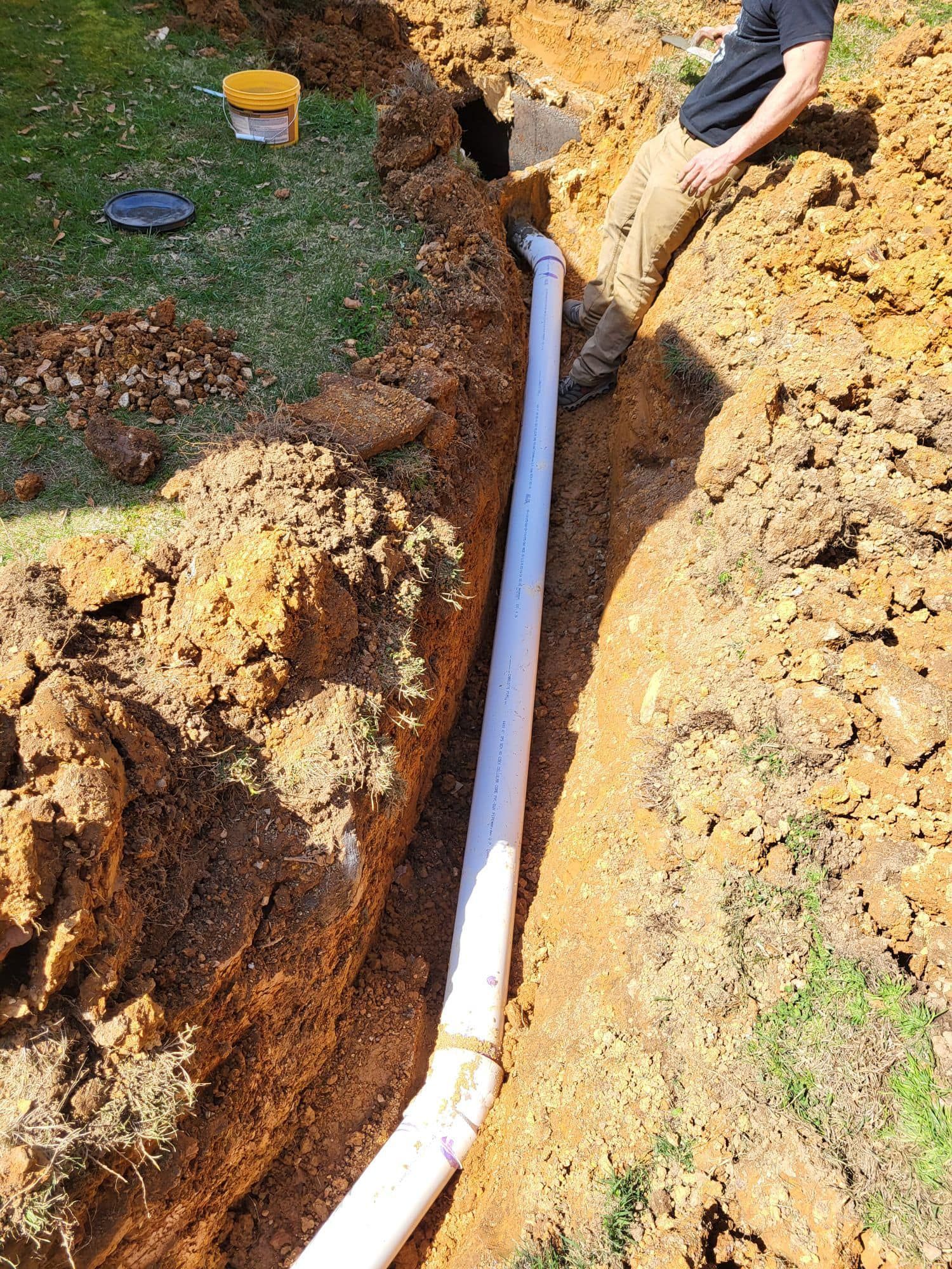 Trench with white PVC pipe installed, person standing nearby. Soil is brown, and grass is green.