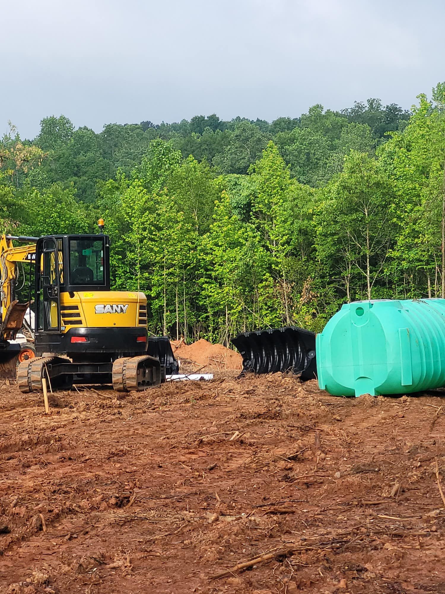A yellow excavator sits on red dirt near a septic tank, with a forest in the background.
