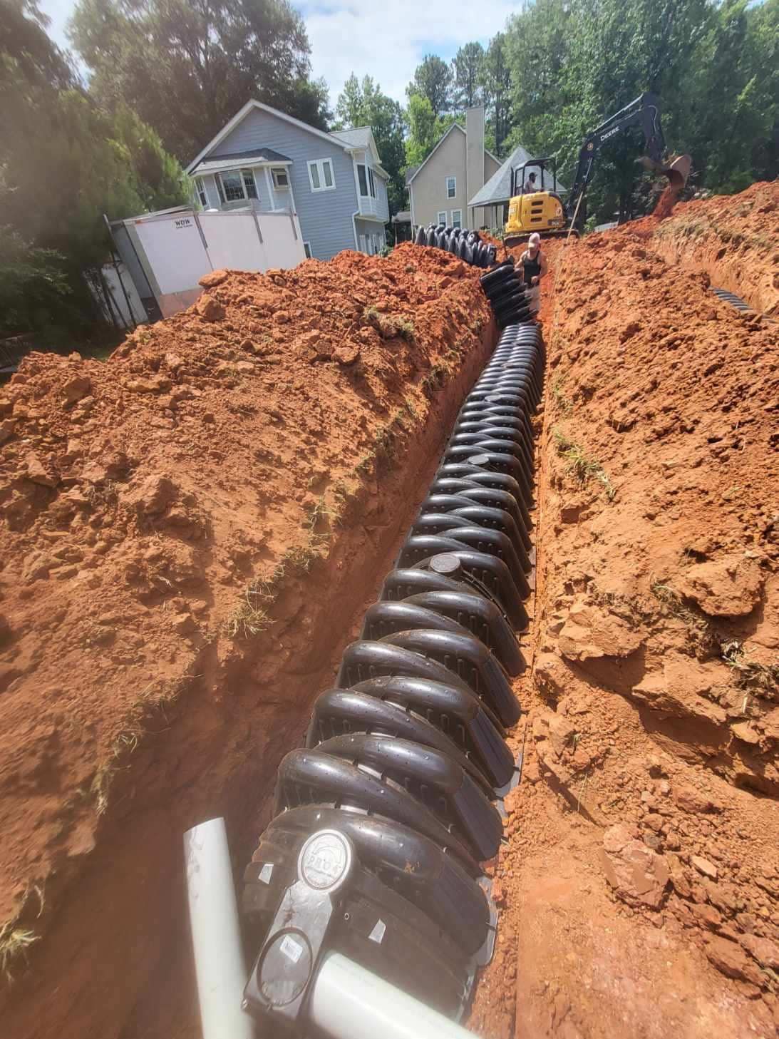 A long trench holds black corrugated drainage pipes. Red soil surrounds the pipes with houses visible in the background.