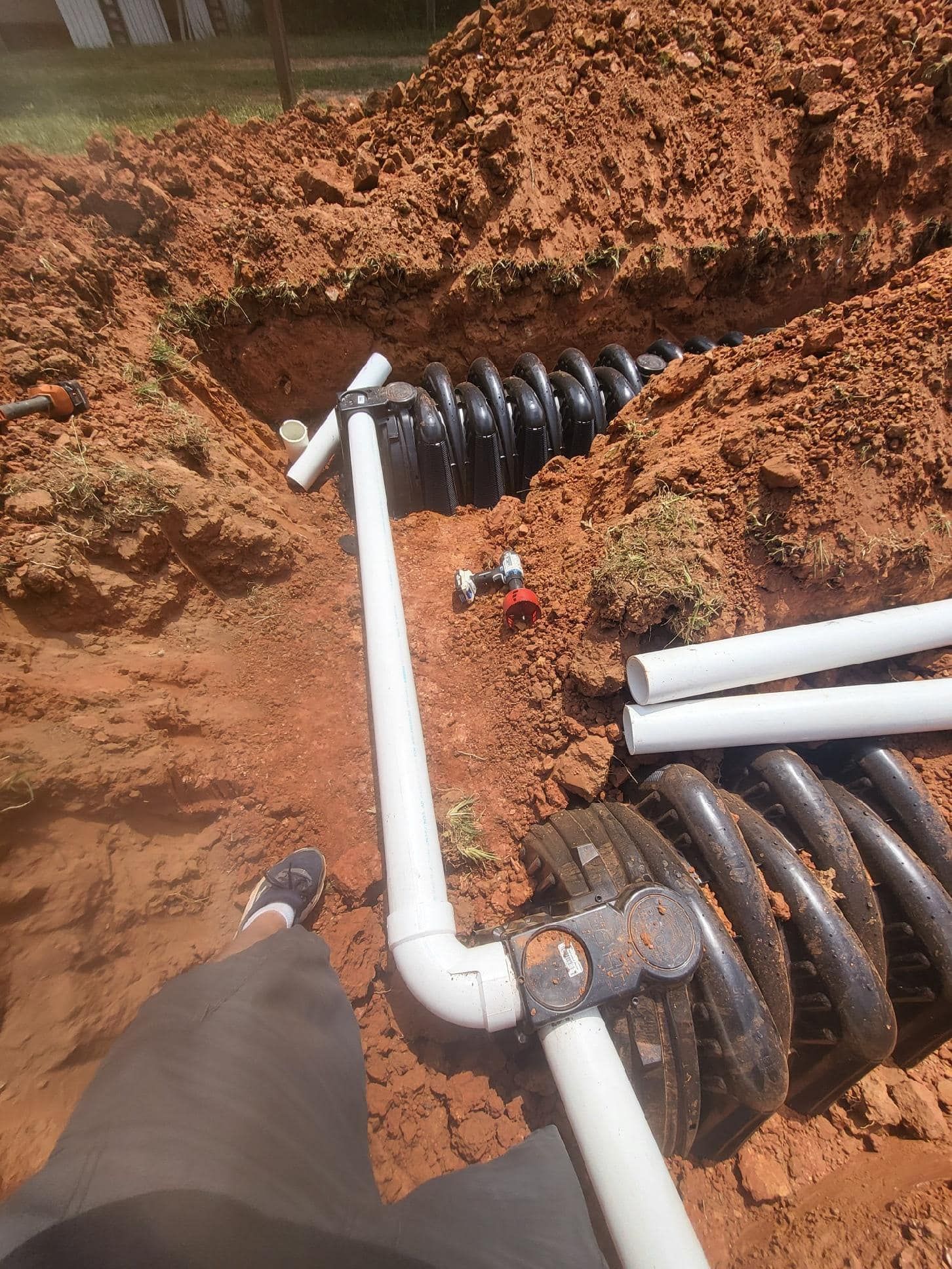 Construction site: trench with black corrugated piping and white PVC pipes. Red clay soil surrounds the work.