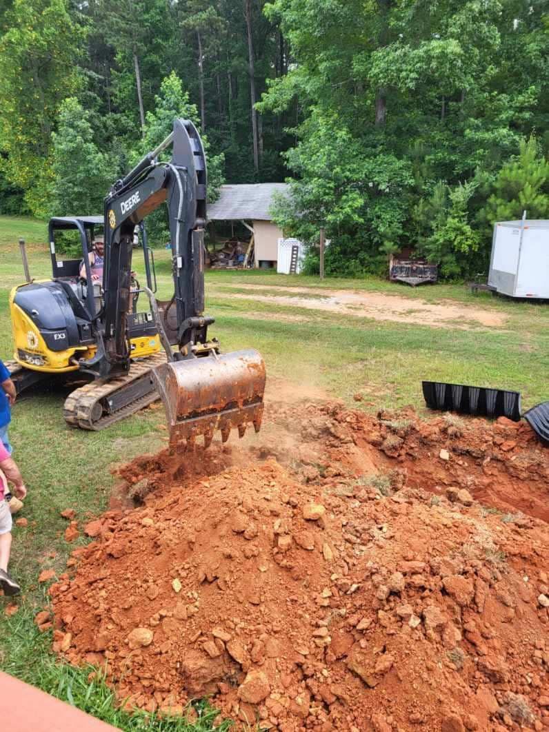 An excavator digs into reddish-brown soil, next to a pile of dirt and black drainage pipes.