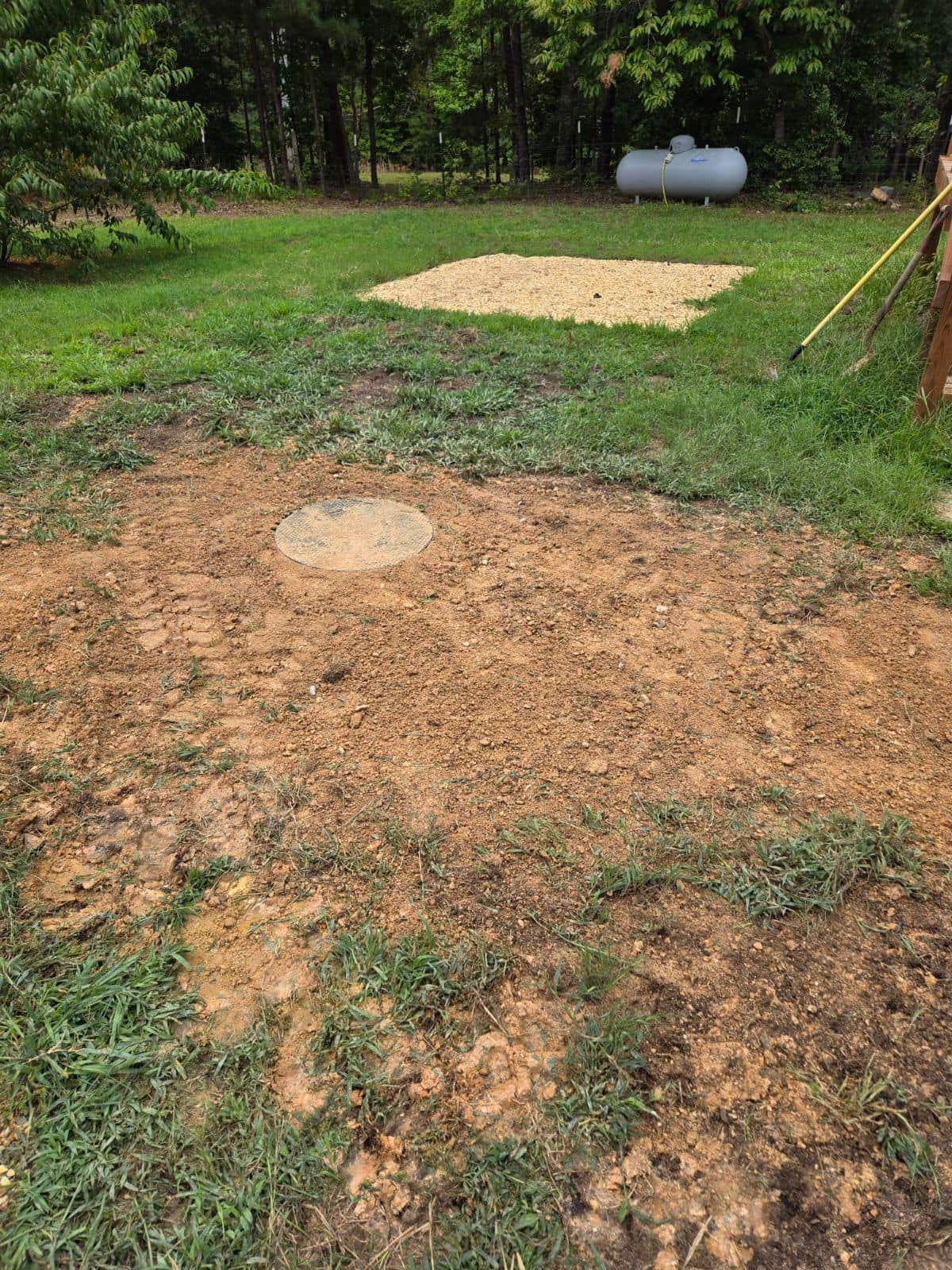 Dirt yard with a circular concrete cover in the foreground, a sand patch, and propane tank in the background.
