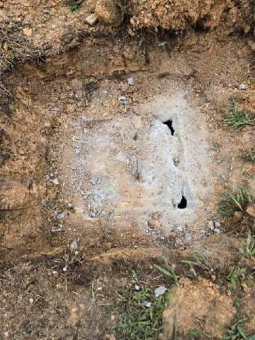 Concrete slab with three holes in the center, partially buried in dirt and surrounded by grass.