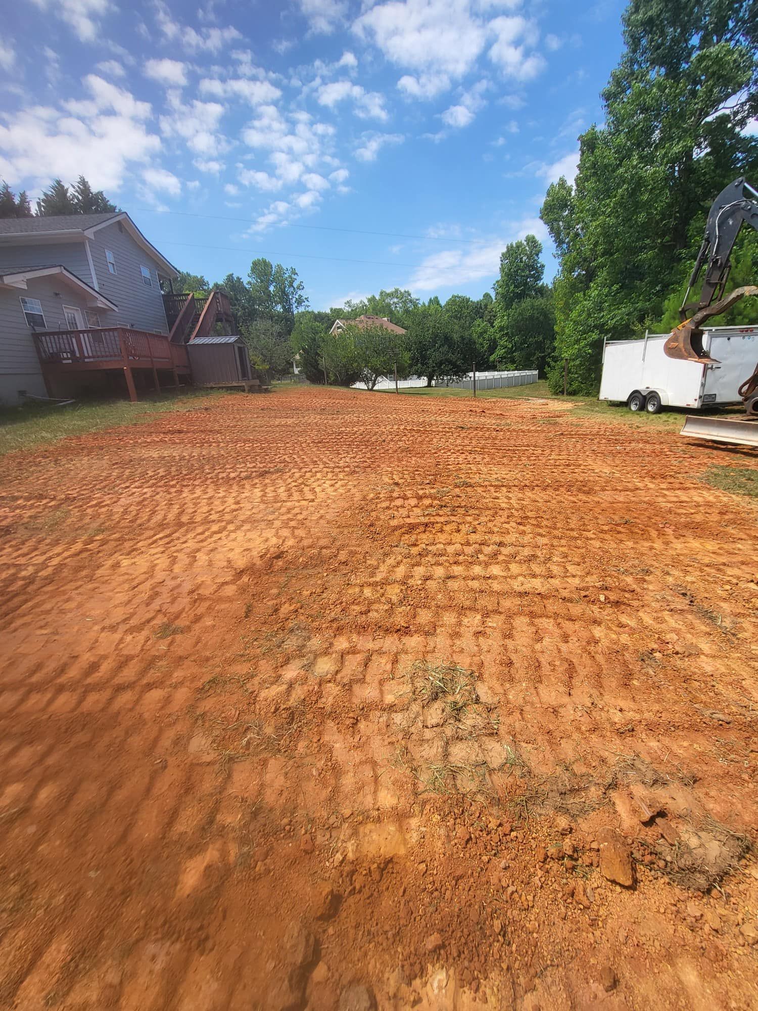 A cleared red dirt lot, possibly for construction, under a blue sky with some trees and a house visible in the background.