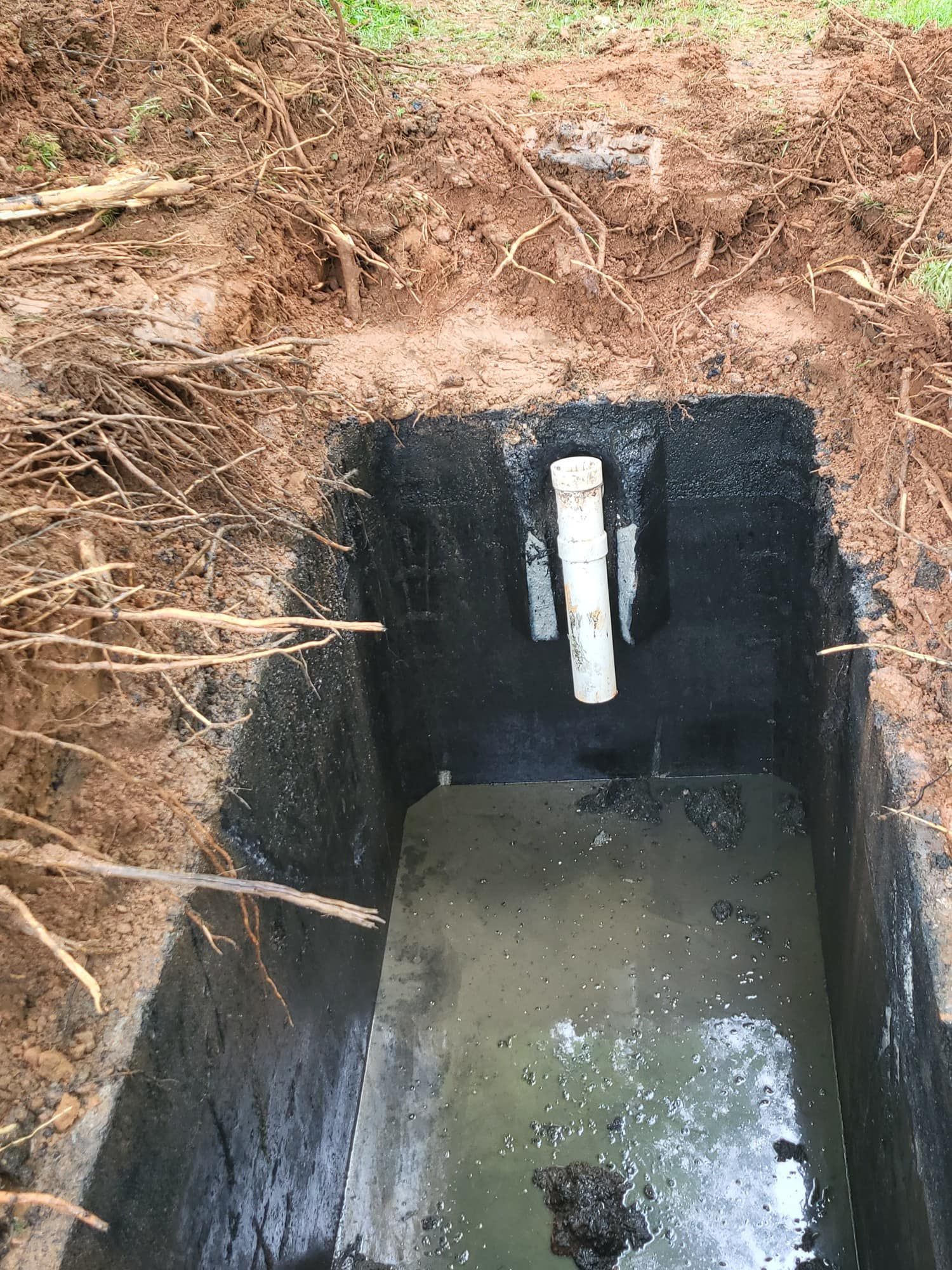 A rectangular septic tank, dug into the earth. The tank has standing liquid and a white PVC pipe.