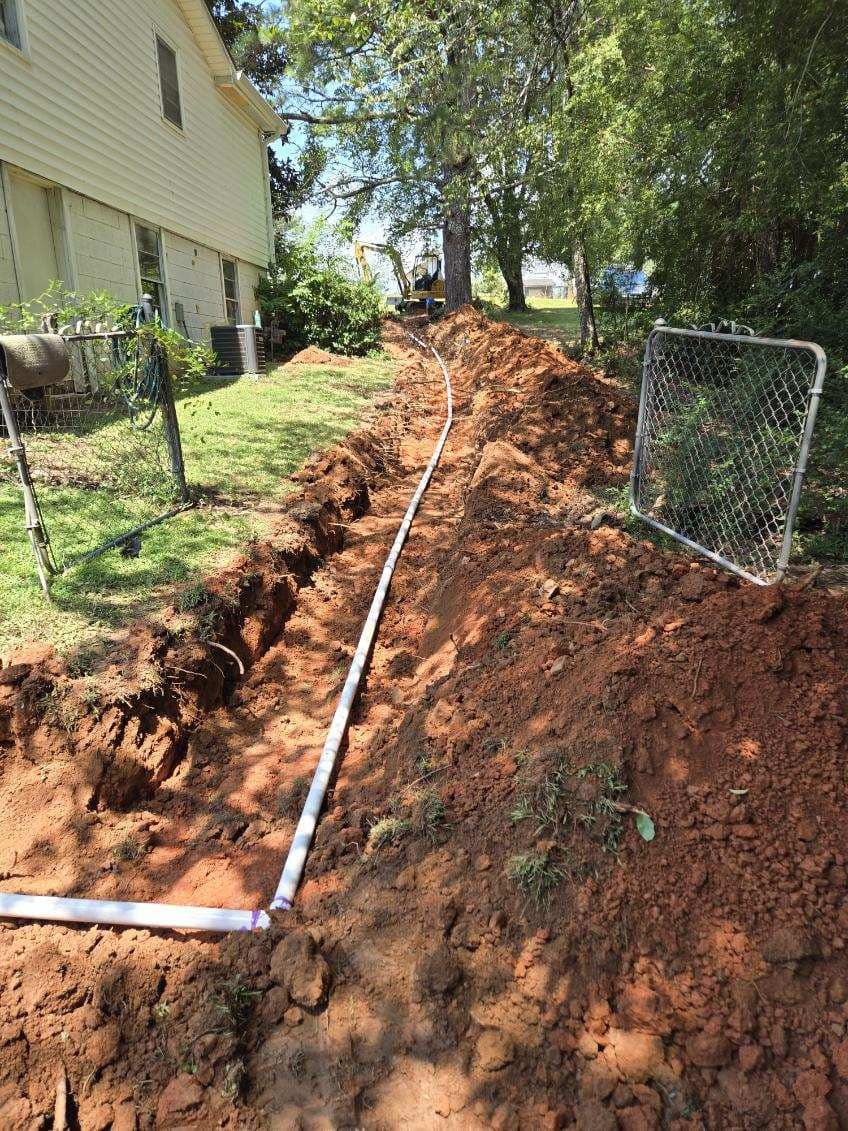 A trench with white pipe running through it in a backyard next to a house. Dirt mounds border the trench.