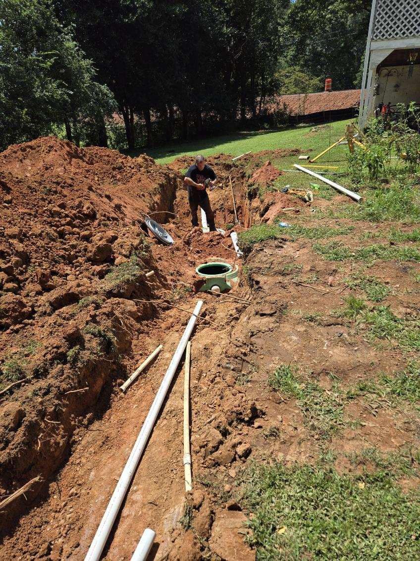 A person works in a long trench, surrounded by dirt and pipes, in a backyard setting.