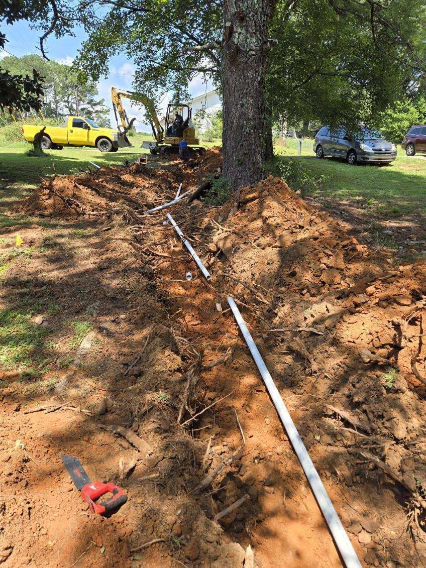A trench dug in a yard with white pipe running through it, near a tree and construction vehicles.