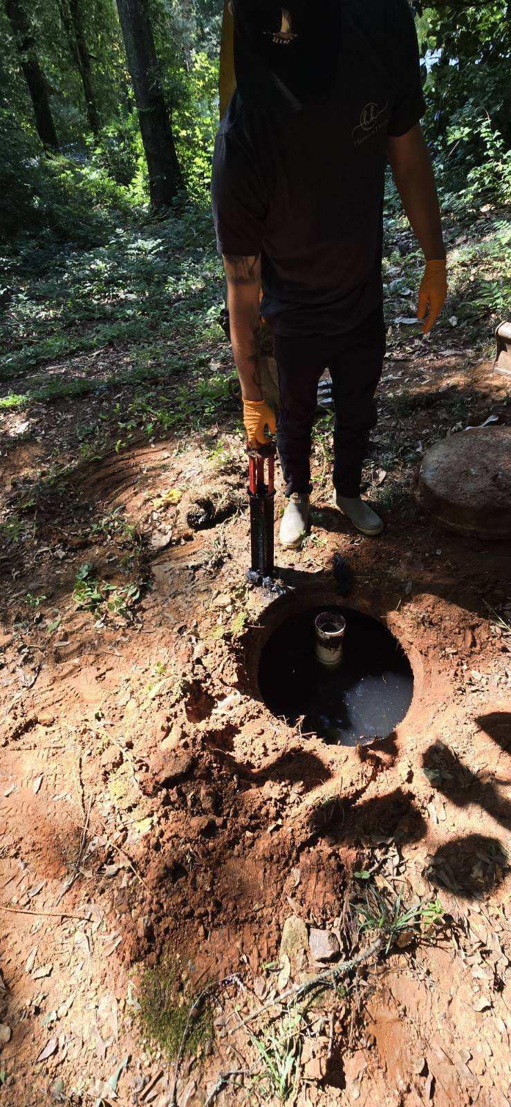 A person is working on a hole in the ground in a wooded area. The hole is surrounded by dirt.