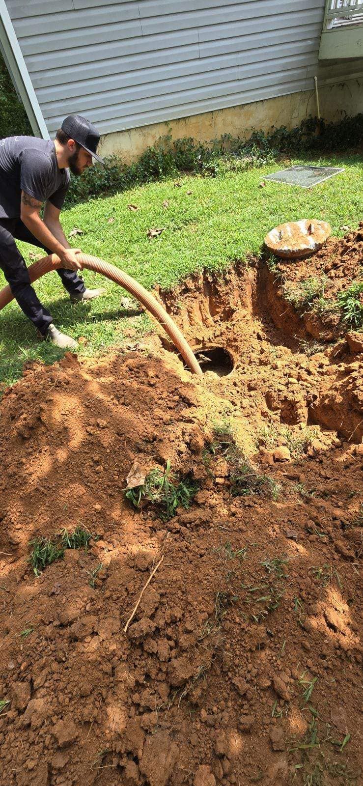 Man using a hose to pump out a septic tank in a backyard. Dirt is piled around the tank.
