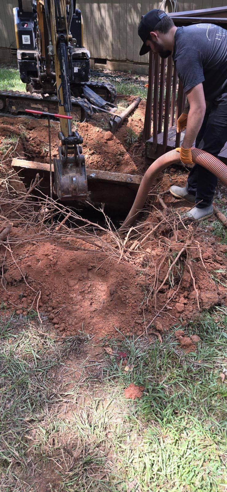 A person uses a hose to remove debris from a hole dug next to a wooden deck. An excavator is nearby.